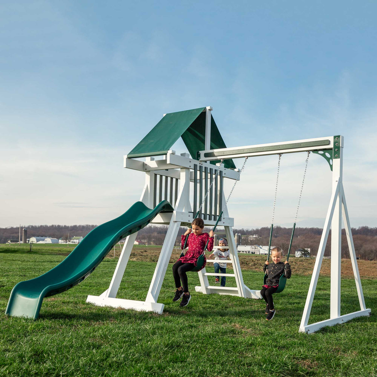 Children swinging on a white vinyl backyard playset with green tarp roof, wave slide and two swings