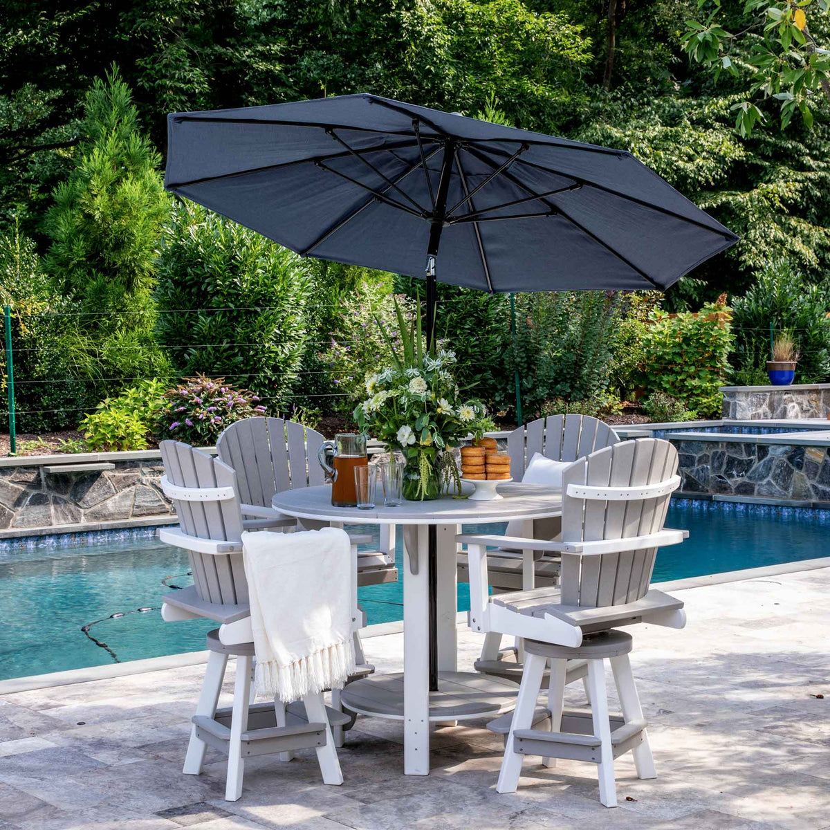 Light gray and white poly counter height patio set with round table, umbrella and four swivel Adirondack chairs beside a pool