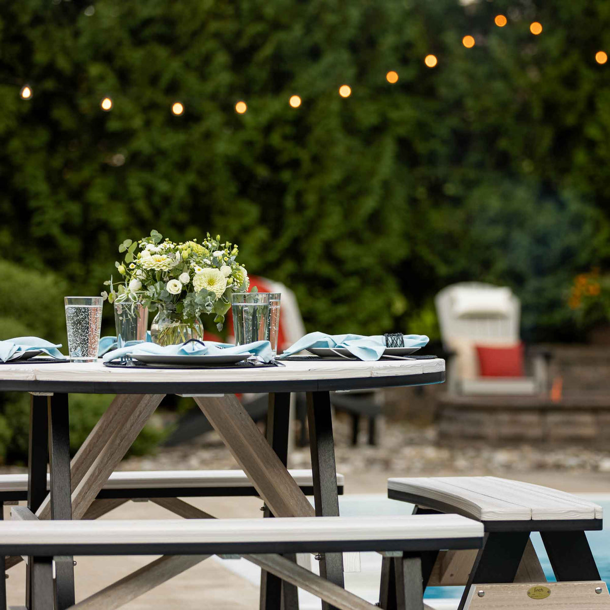 Table setting on a round weatherwood poly picnic table with flowers and blue napkins under patio string lights