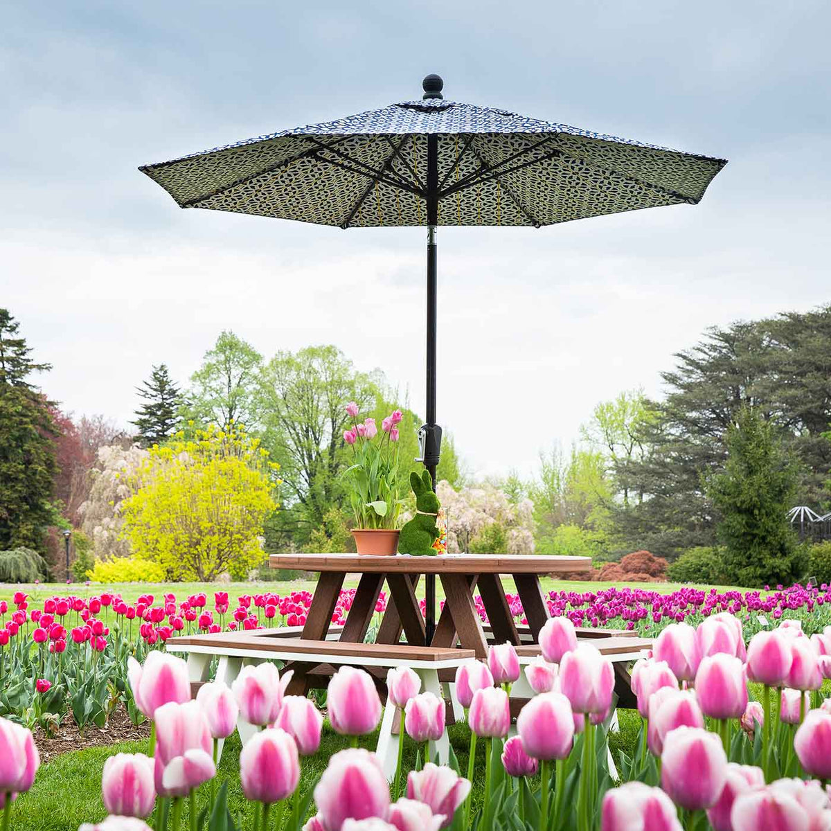 Round poly picnic table with attached benches and a patterned umbrella in a field of pink tulips