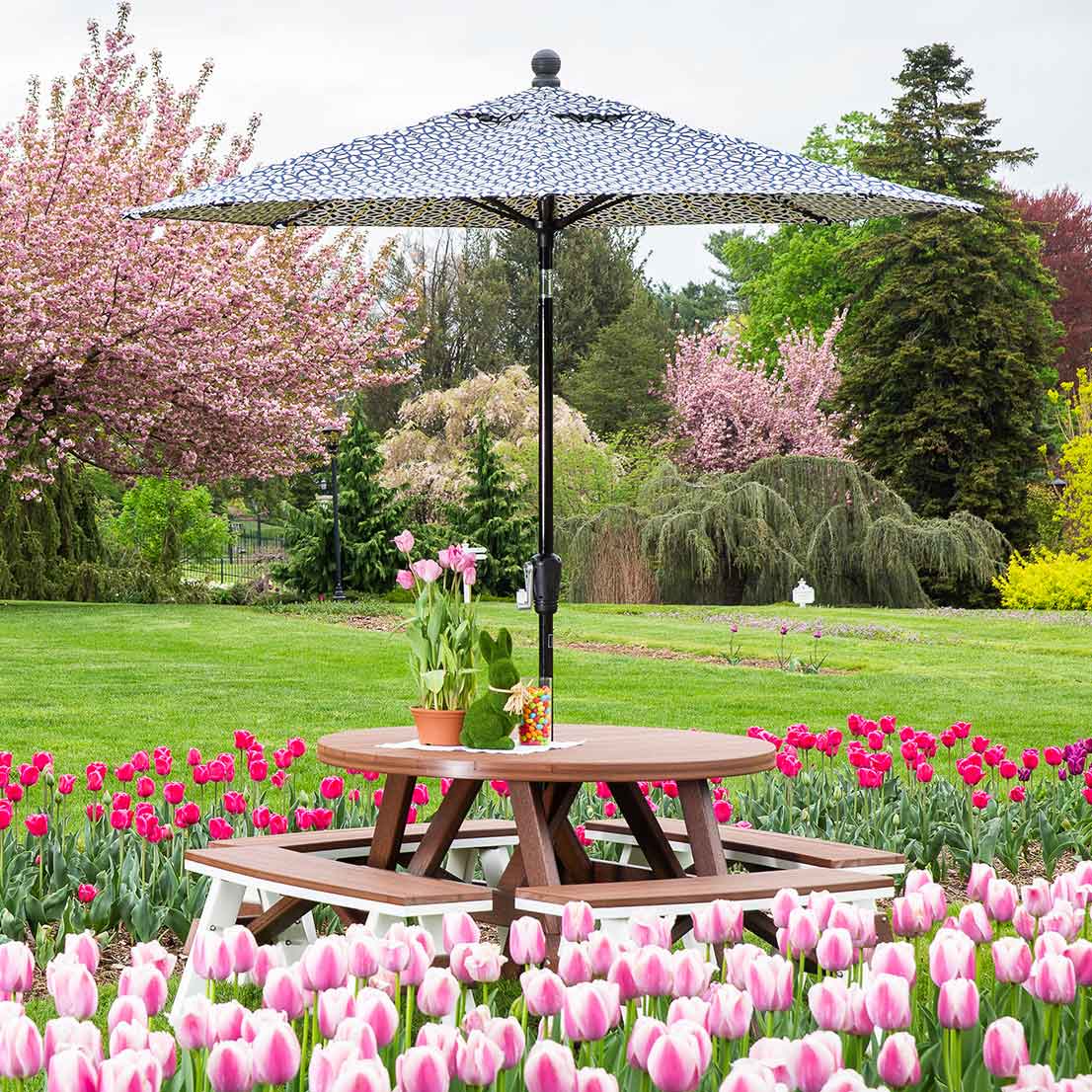 Brown and white round poly picnic table with wraparound benches and a printed umbrella among blooming tulips
