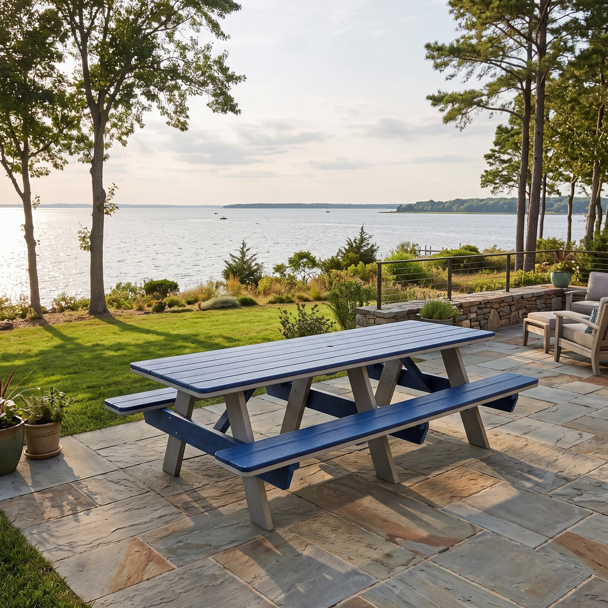 A blue and gray outdoor picnic table with attached benches on a stone patio overlooking a lake, with trees and a lawn in the background.