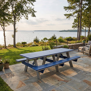 A blue and gray outdoor picnic table with attached benches on a stone patio overlooking a lake, with trees and a lawn in the background.