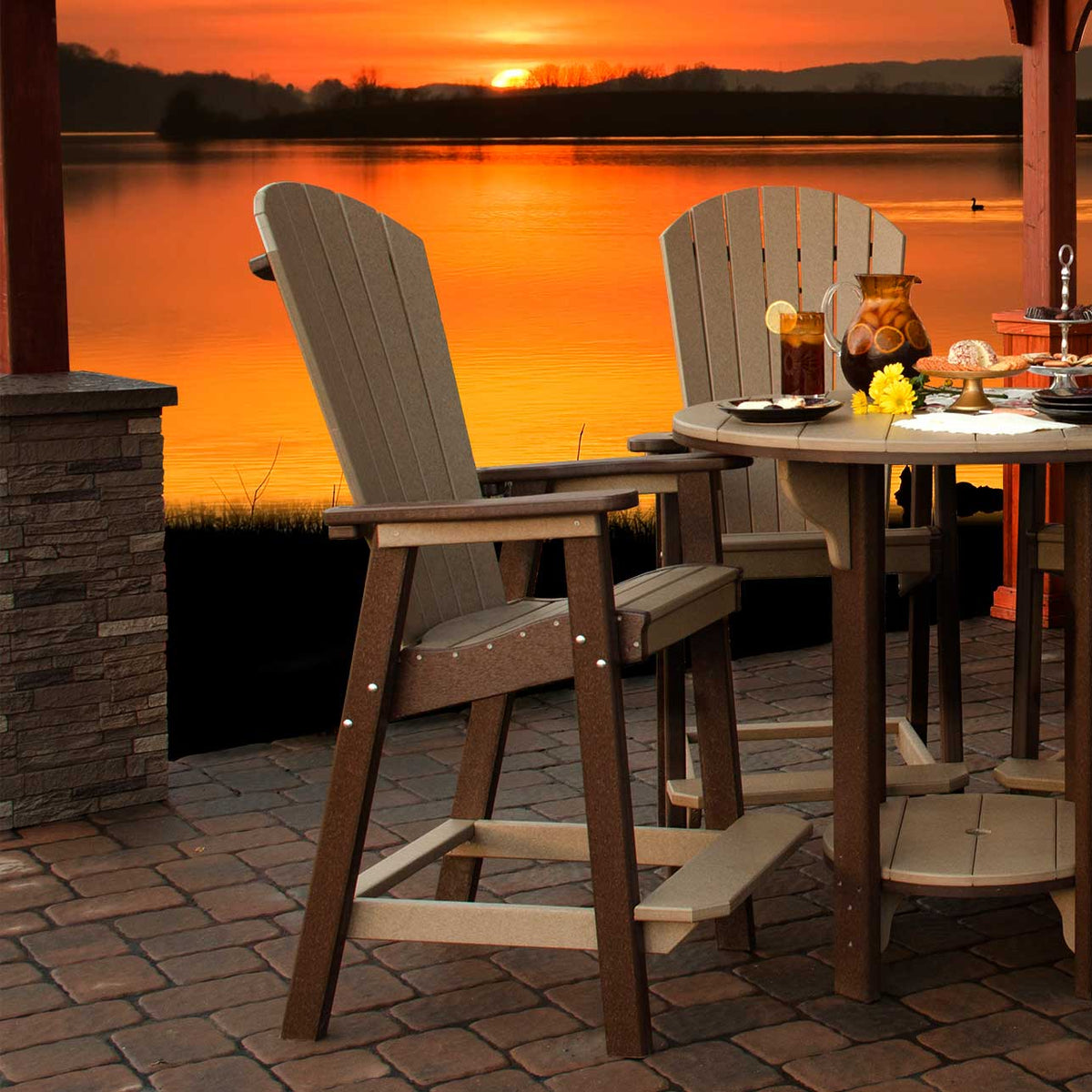 Two weatherwood bar height Adirondack chairs at a round pub table on a paver patio at sunset over the lake