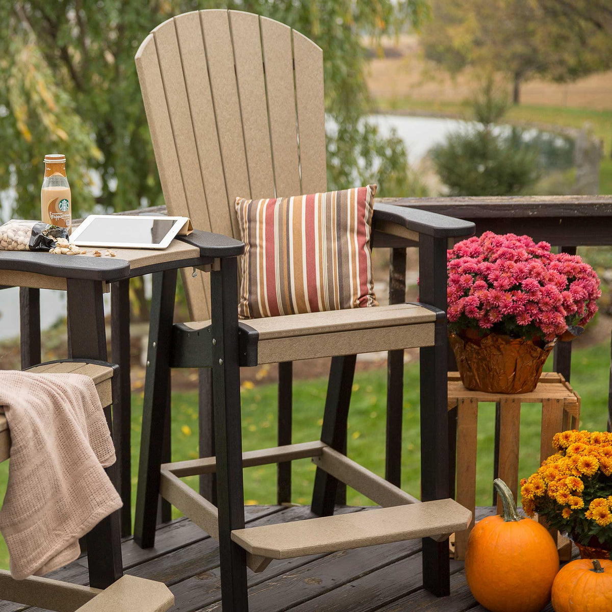 Weatherwood and black tall Adirondack chair on a deck with a striped pillow, mums and pumpkins in autumn