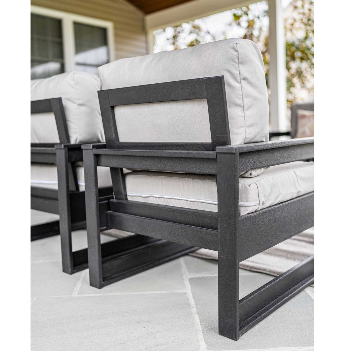 Side view of black poly club chairs with open square frames and thick gray cushions on a porch