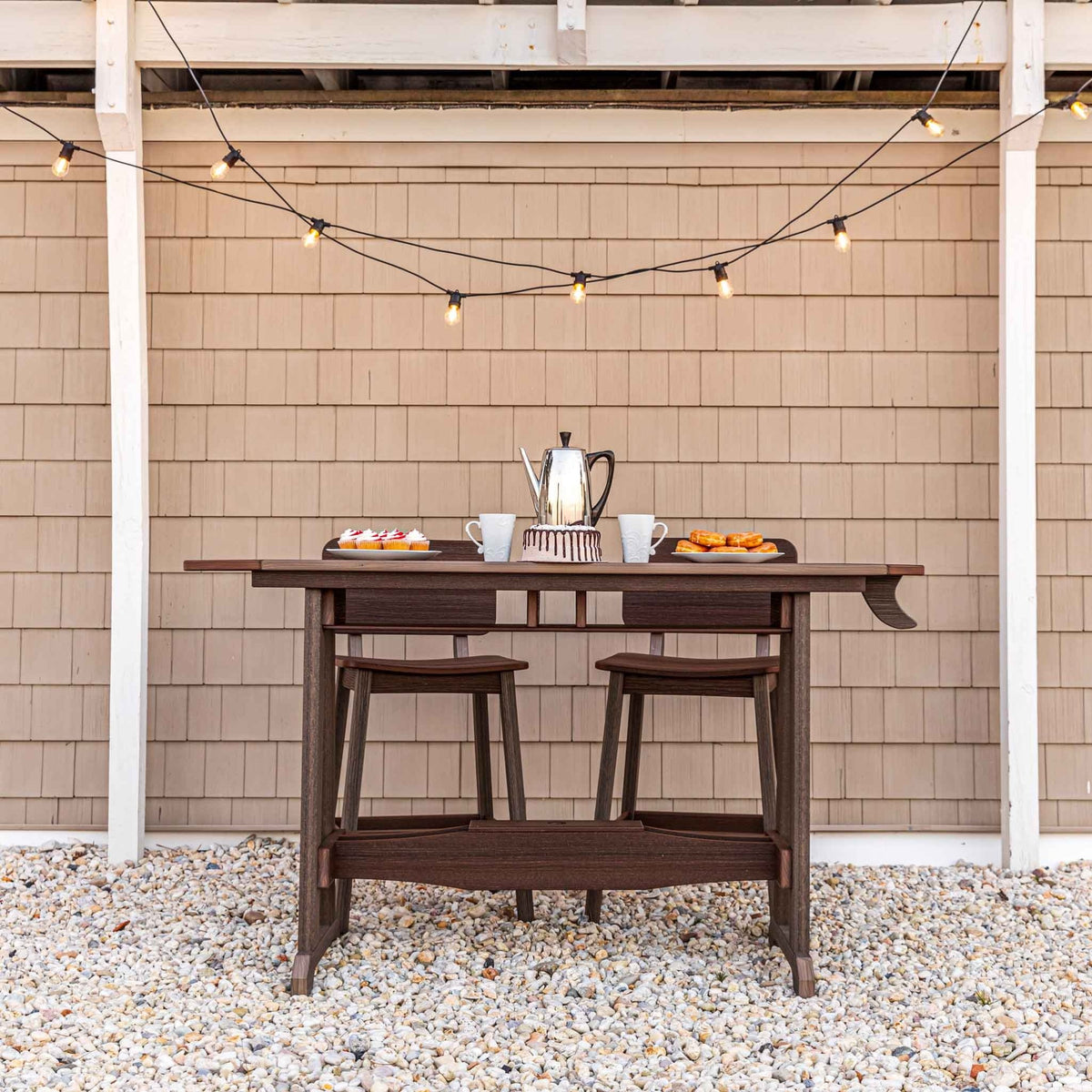 Dark brown recycled poly counter table with two backless stools tucked underneath on a gravel patio with string lights