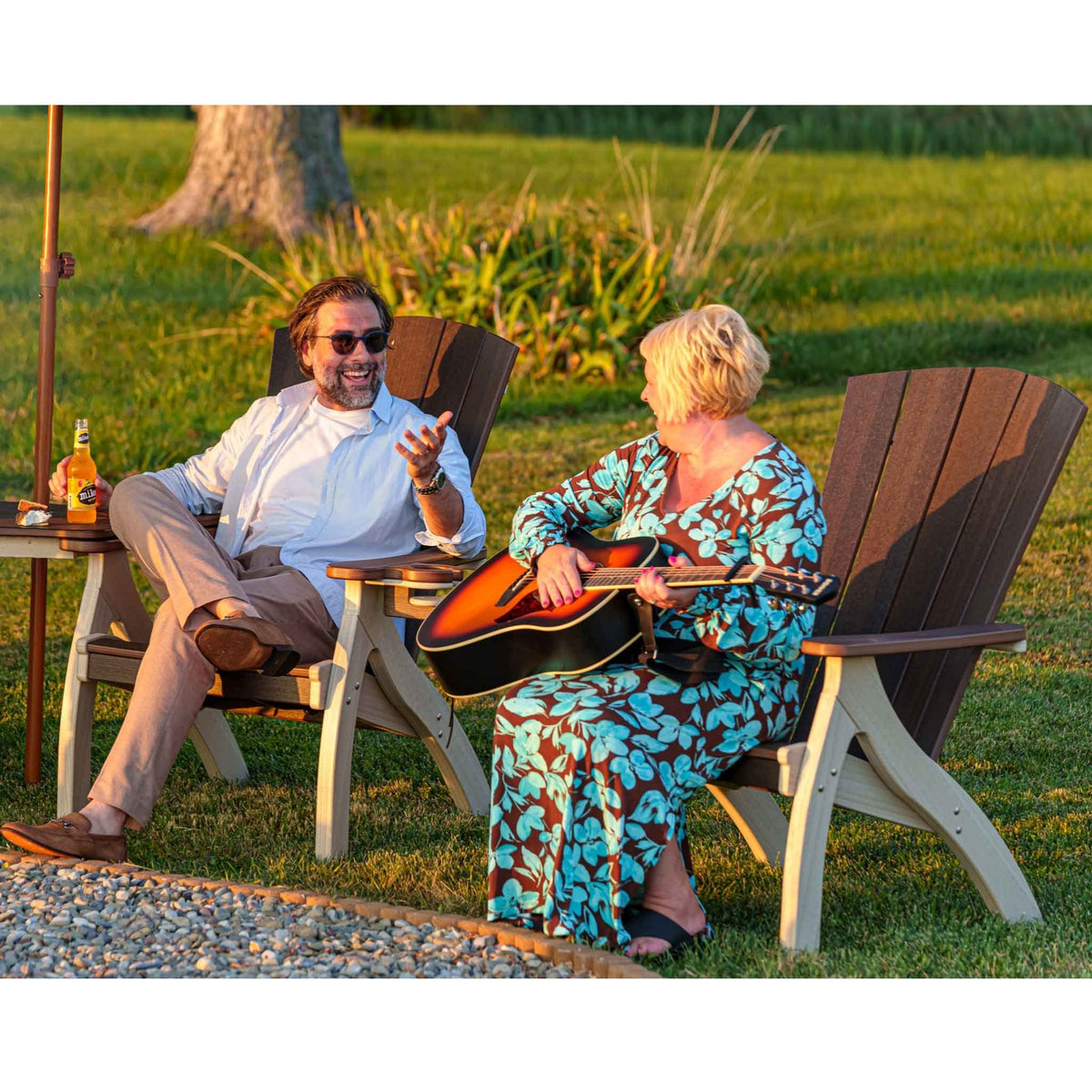 Couple relaxing in brown and weatherwood poly siesta chairs on a lawn at sunset, one playing an acoustic guitar