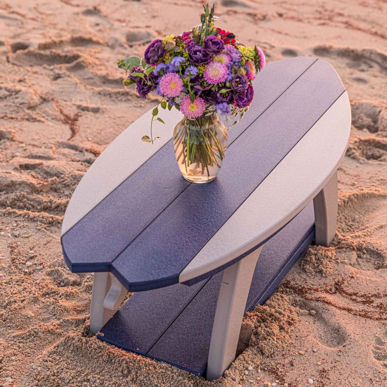 Navy and gray surfboard shaped coffee table with a lower shelf and a vase of flowers on the sand