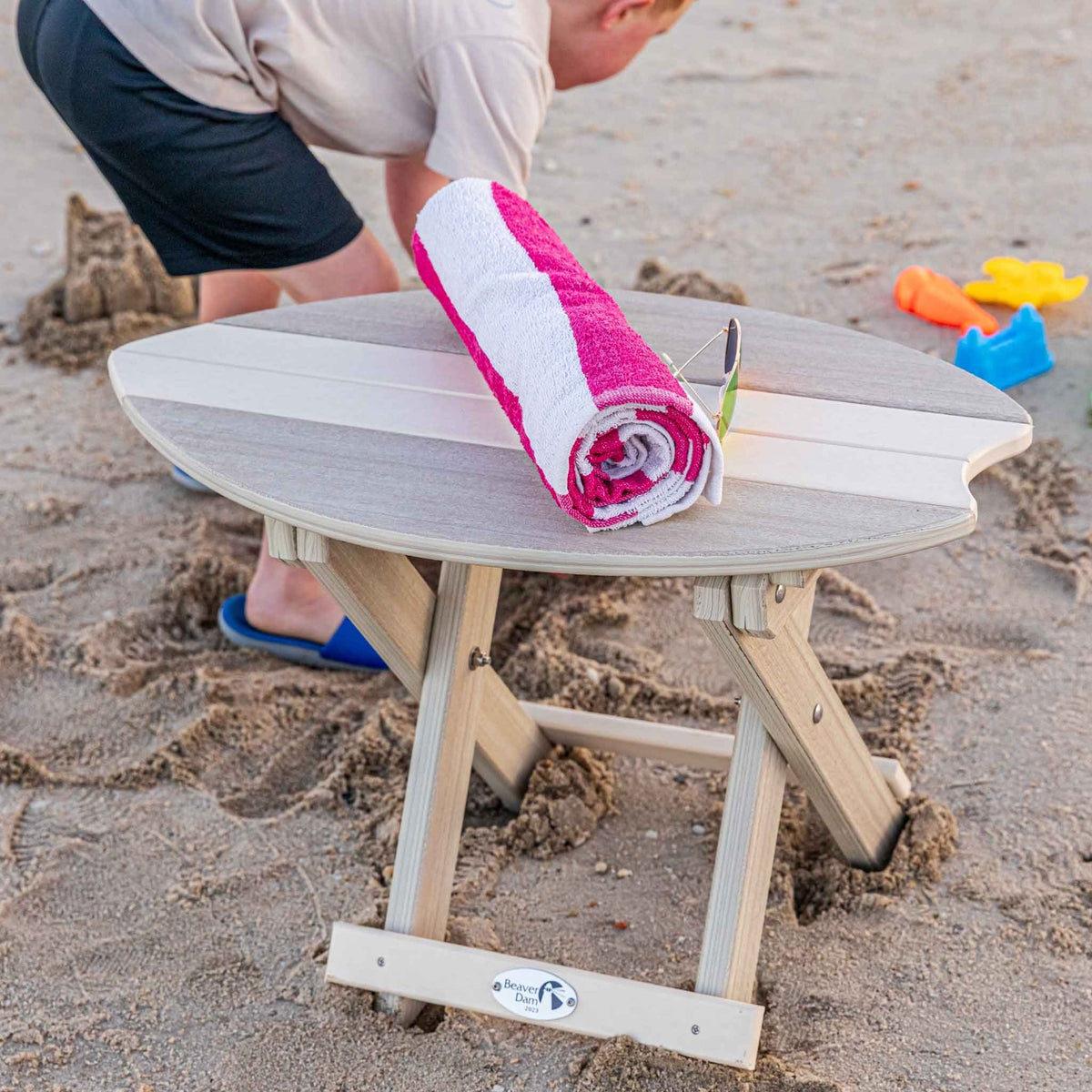 Child reaching for a rolled pink striped towel on a gray surfboard shaped folding beach table