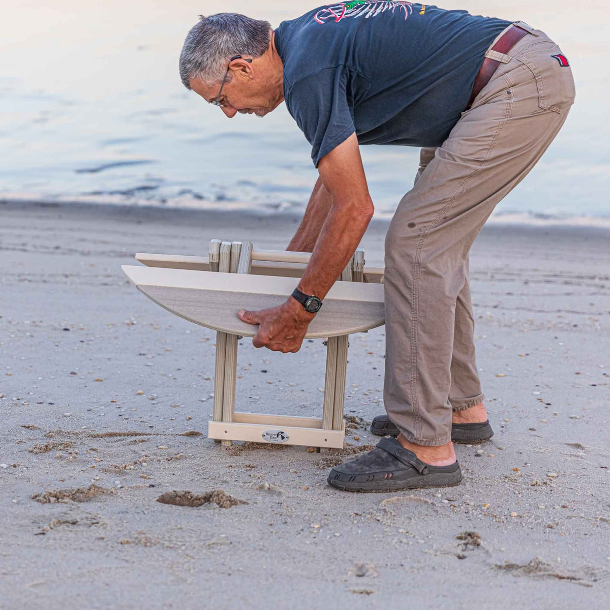 Man setting up a weatherwood folding surfboard shaped end table on the beach at dusk