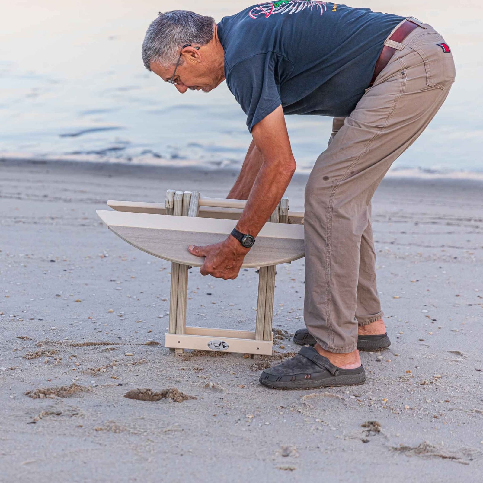 Man setting up a weatherwood folding surfboard shaped end table on the beach at dusk