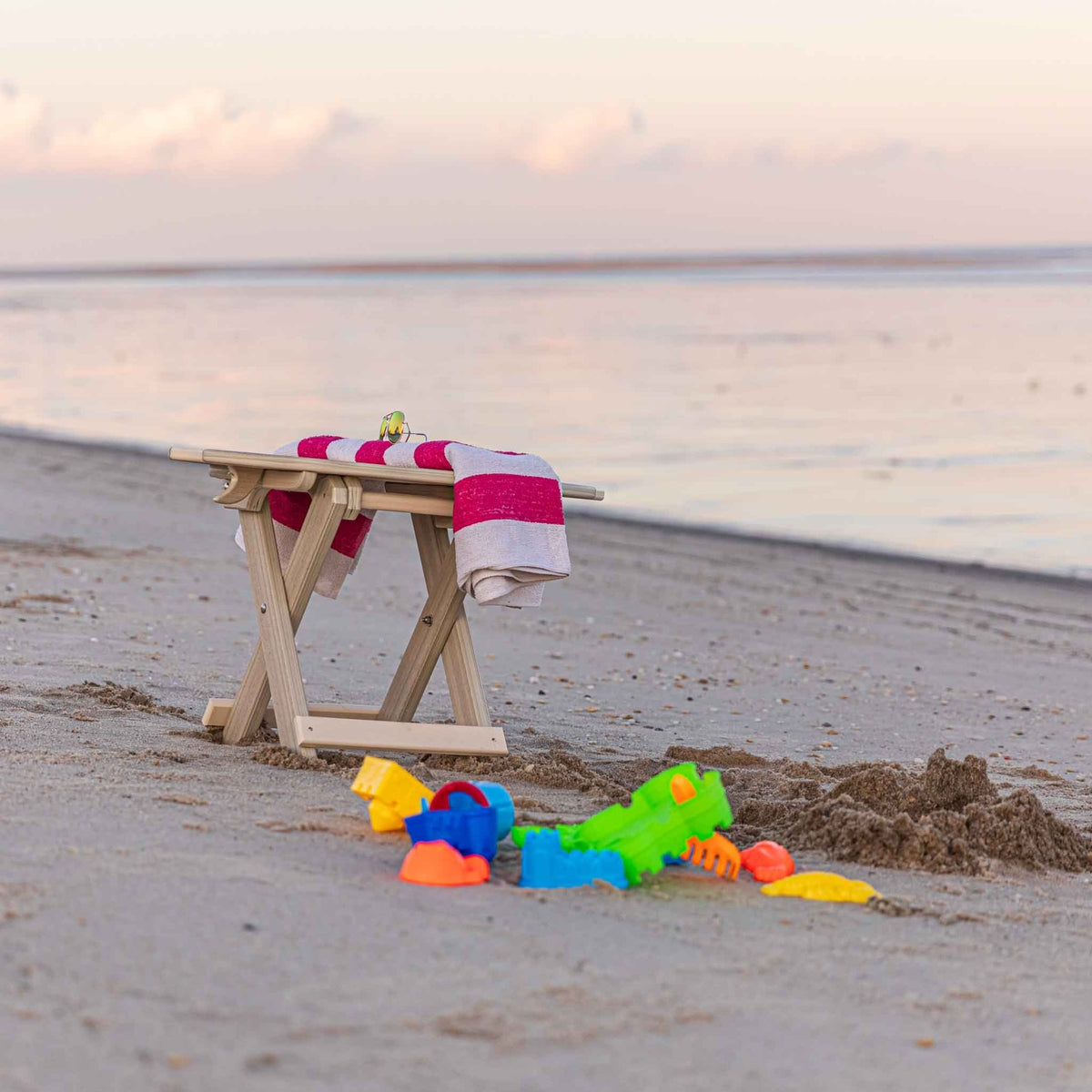 Weatherwood folding surfboard side table with a striped towel draped over it and sand toys nearby