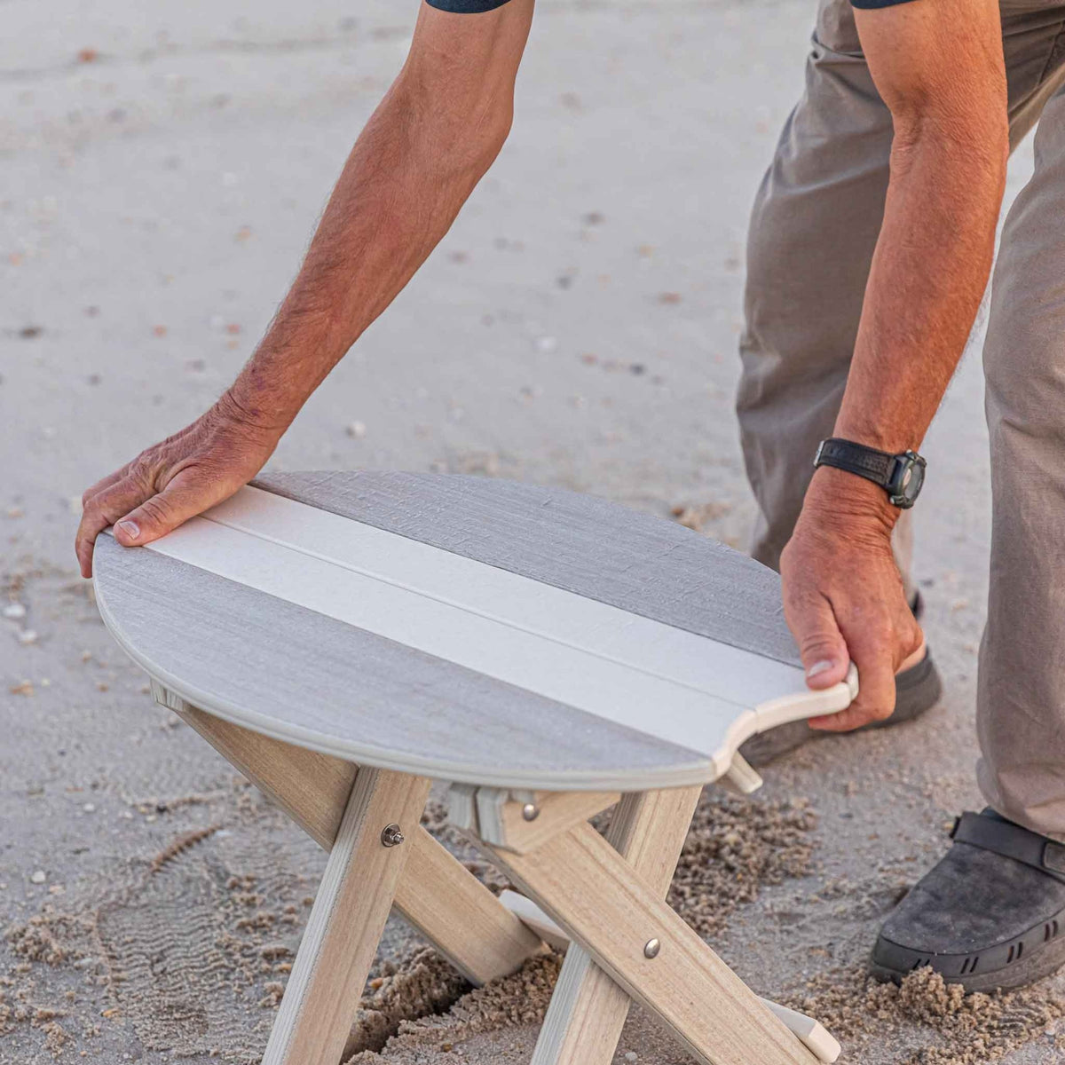 Hands opening the weatherwood surfboard shaped folding side table with X frame legs on the sand