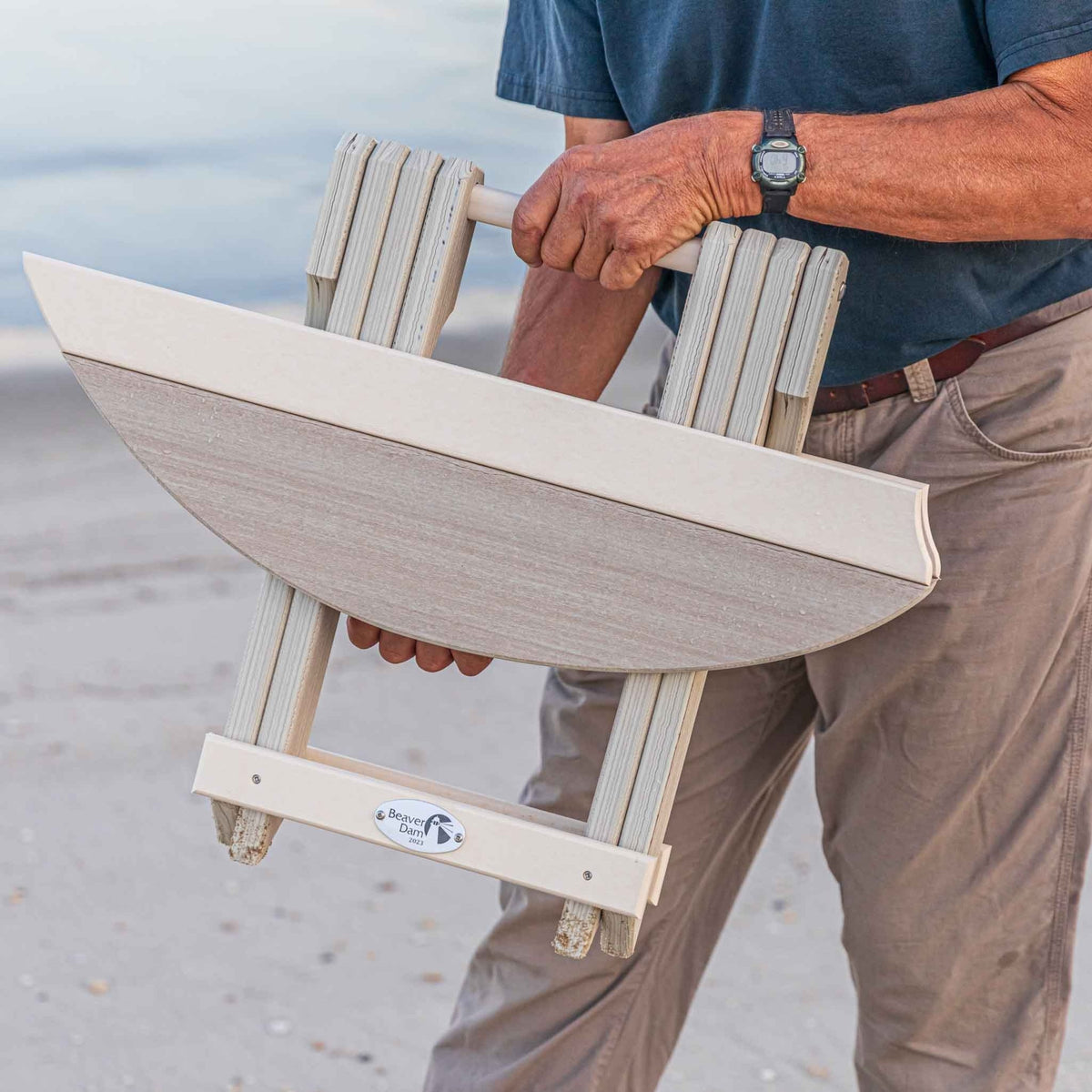 Person carrying the folded driftwood poly surfboard end table by its built-in handle on the sand