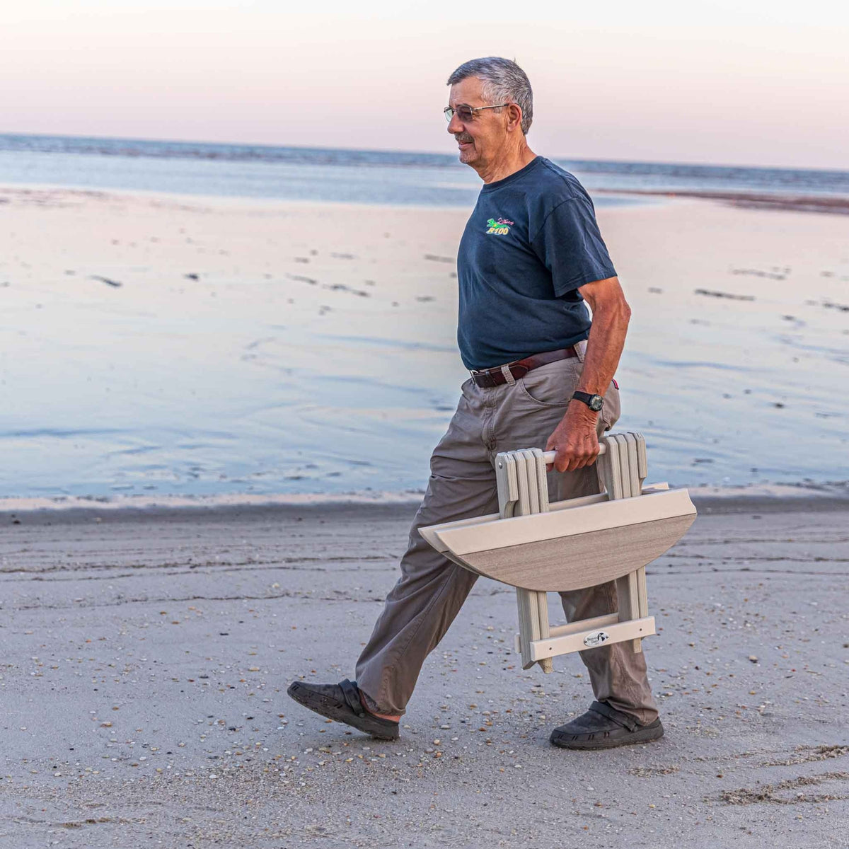 Man walking along the shore carrying the folded driftwood surfboard end table at his side