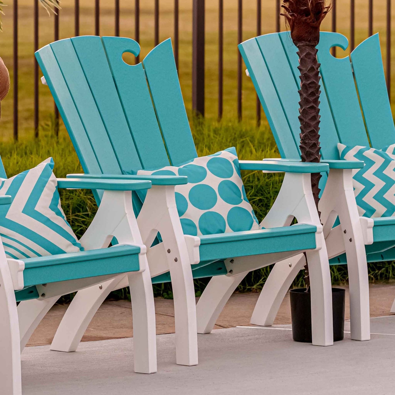 Row of turquoise and white wave back patio chairs with throw pillows beside a pool and palm tree