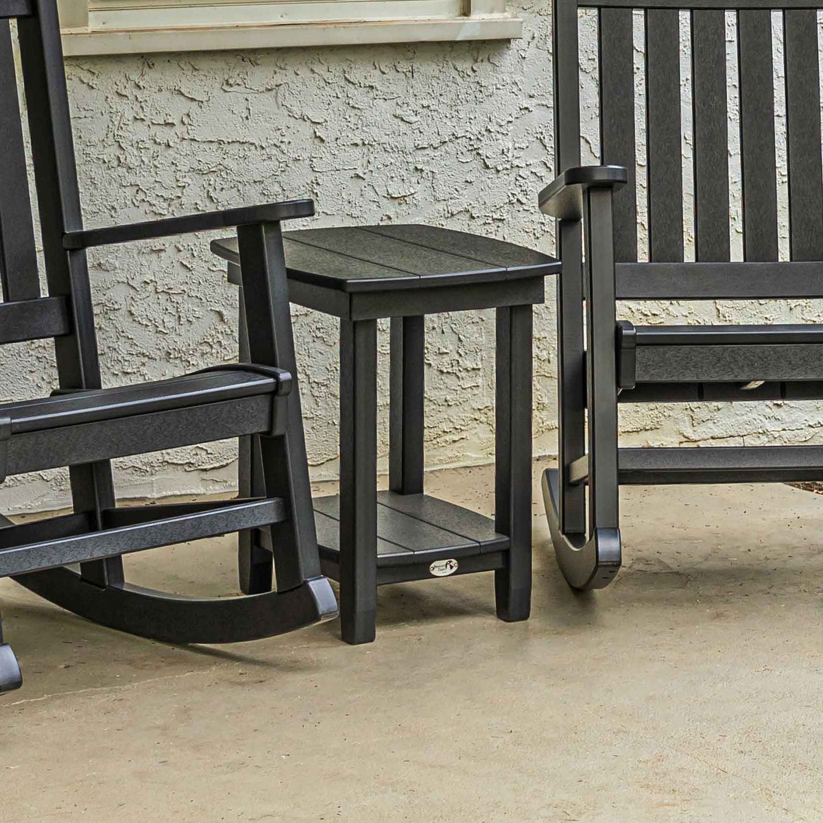 Black poly end table with a lower shelf sitting between two black porch rockers on a concrete patio
