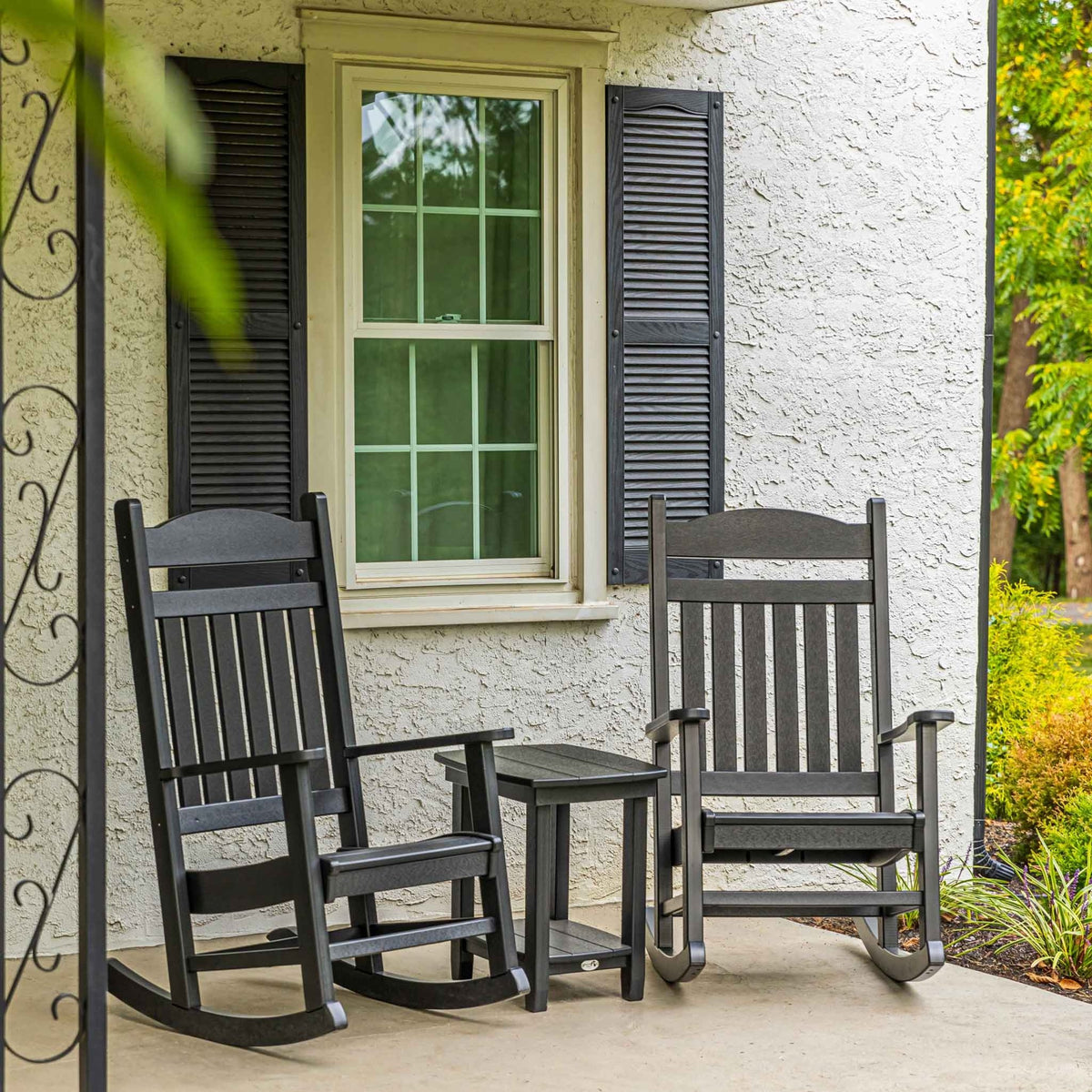 Pair of black slat back rocking chairs and a side table on a covered front porch with greenery
