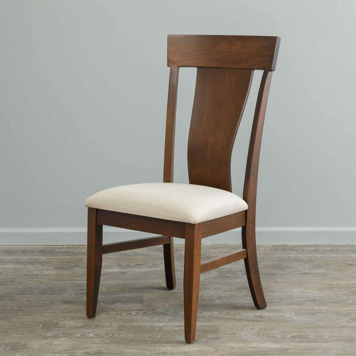 Walnut toned dining chair with a curved hourglass back panel and cream linen upholstered seat, standing on gray wood flooring