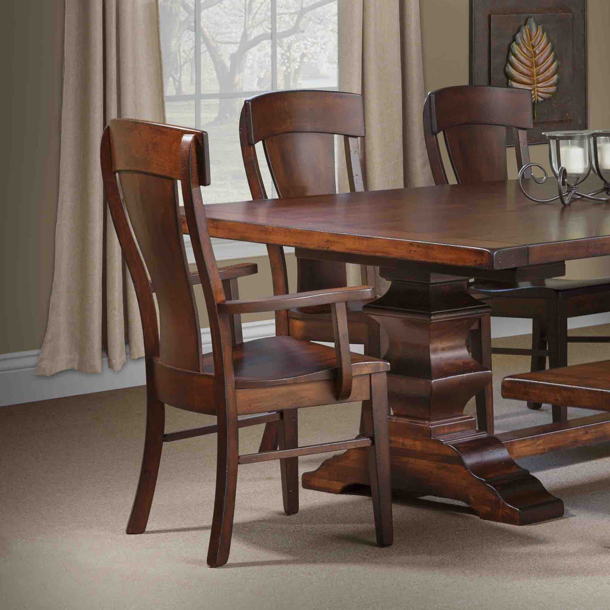 Cherry finished dining arm chair with a curved splat back and shaped arms, pulled up to a dark pedestal dining table by a draped window
