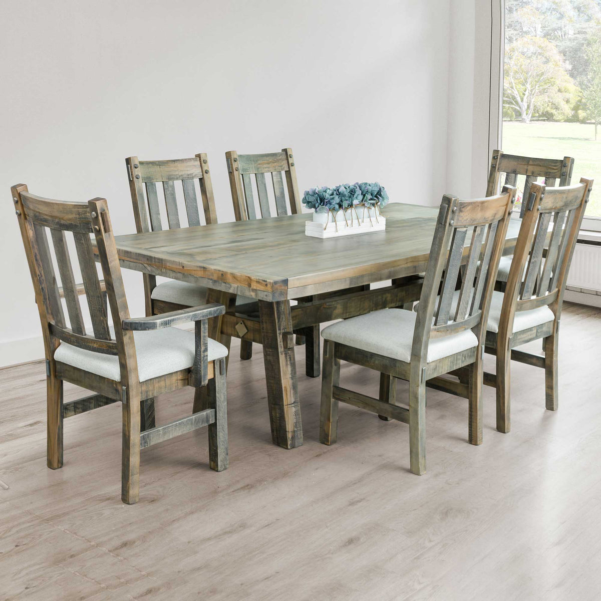 Weathered gray trestle dining table with six slat back chairs upholstered in light fabric by a window