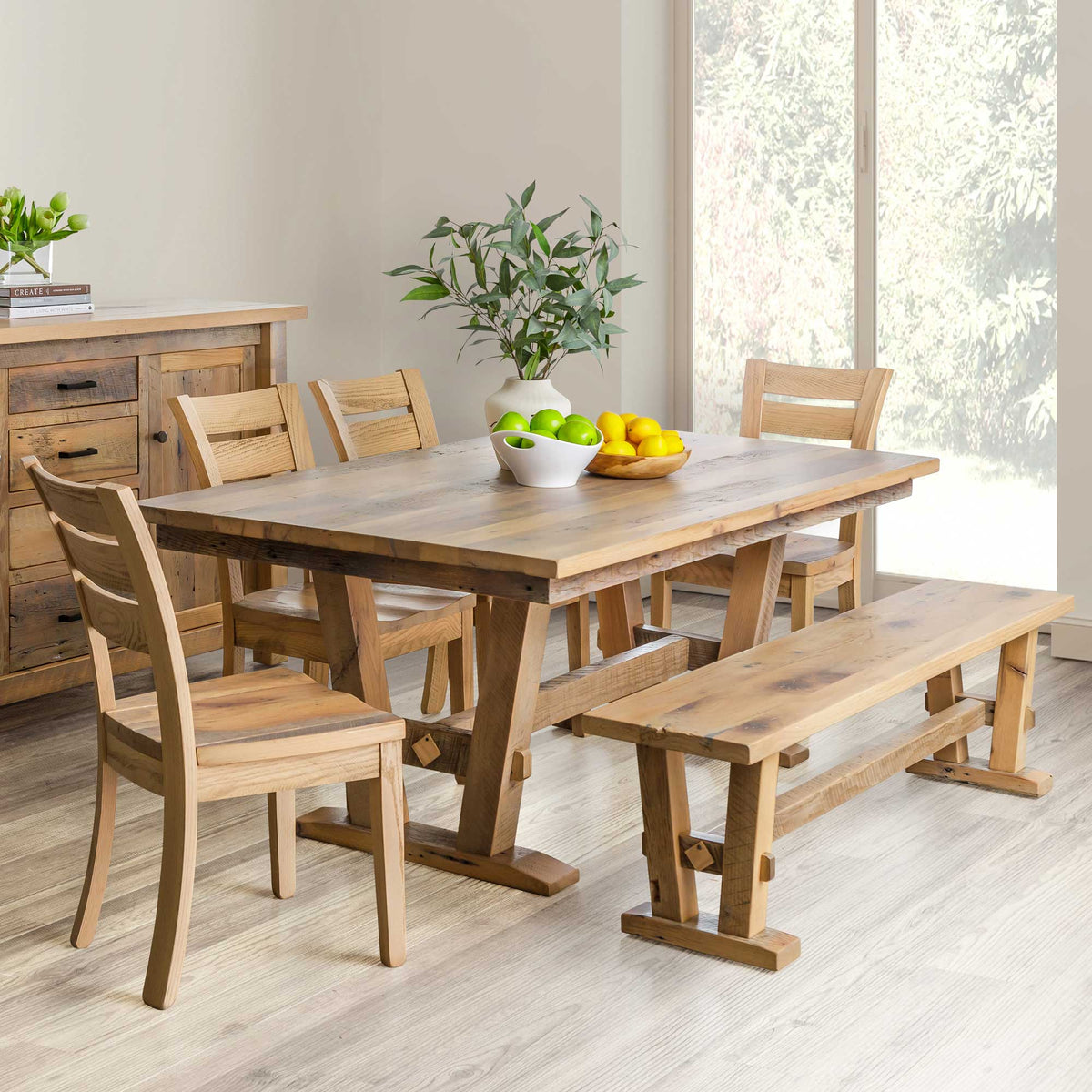 Natural finish reclaimed wood trestle dining table set with four ladder back chairs and a matching bench in a bright farmhouse dining room