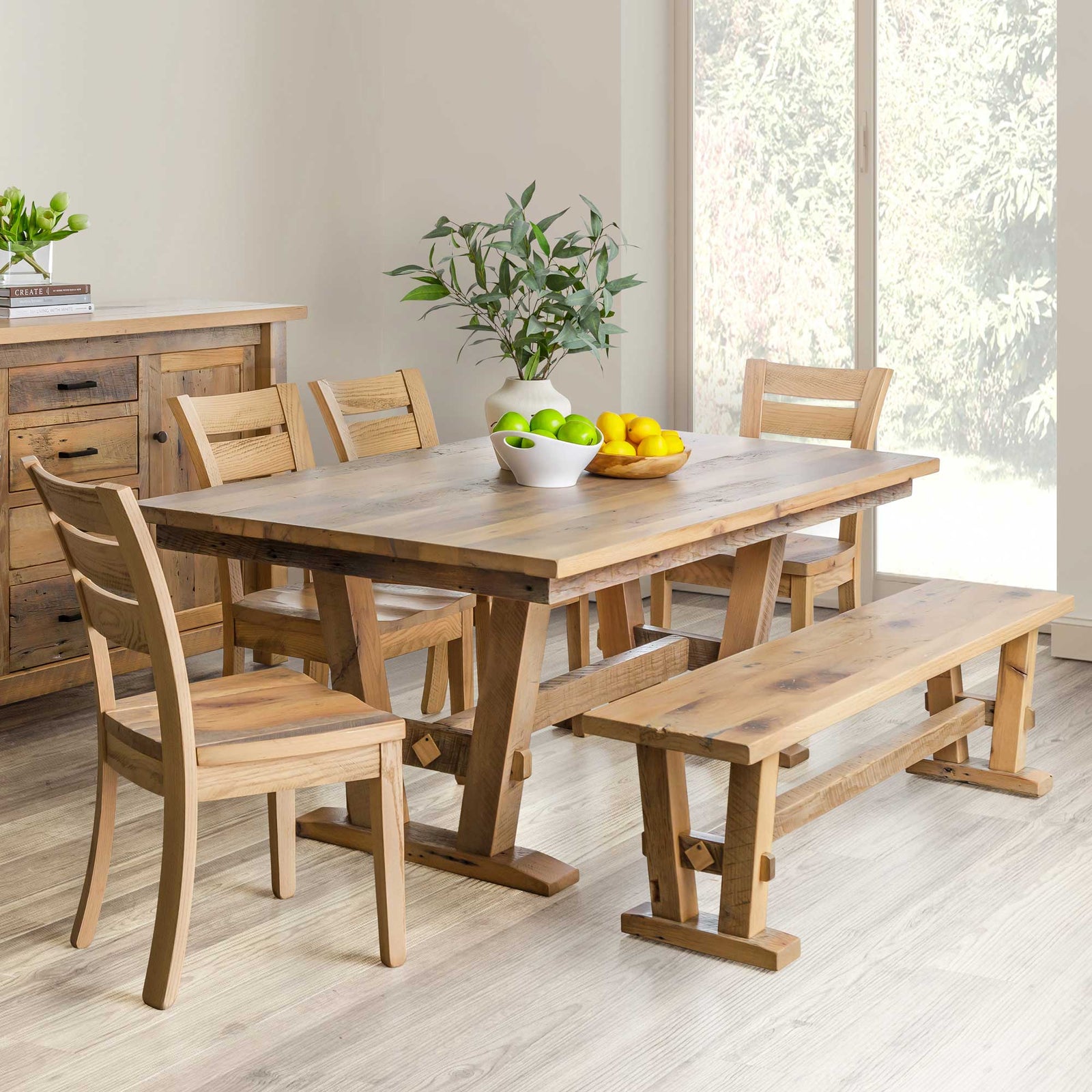 Natural finish reclaimed wood trestle dining table set with four ladder back chairs and a matching bench in a bright farmhouse dining room