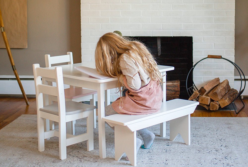 Girl in pink overalls sitting on a white kid's bench at a small white play table in a living room with a fireplace