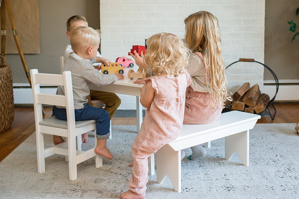 Four children playing with wooden toy cars at a white kid's table with a matching bench and chairs by a brick fireplace
