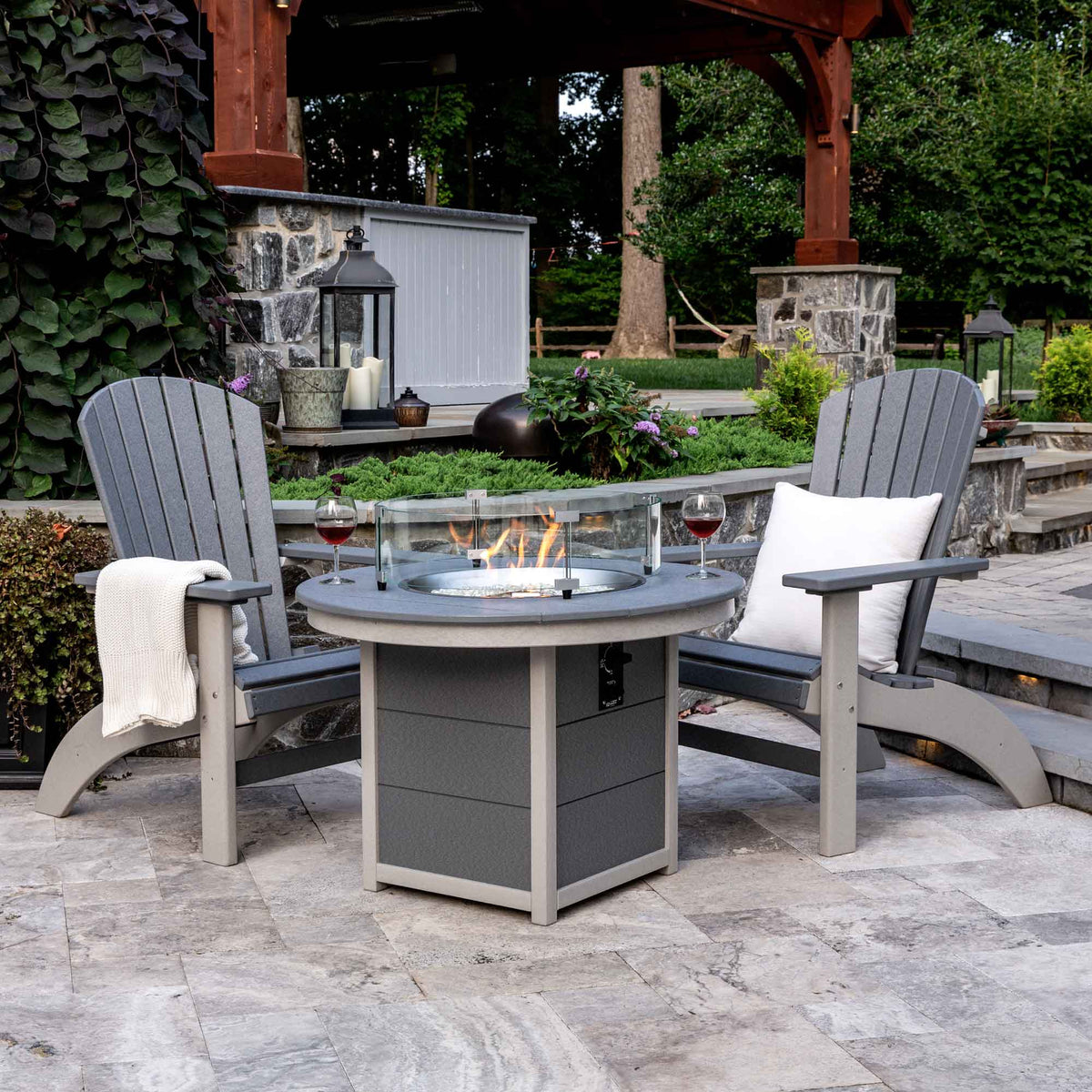 Two gray poly Adirondack chairs facing a lit round poly fire pit table with glass wind guard on a stone patio