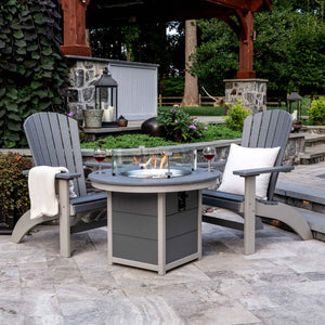 Two gray poly Adirondack chairs facing a lit round poly fire pit table with glass wind guard on a stone patio
