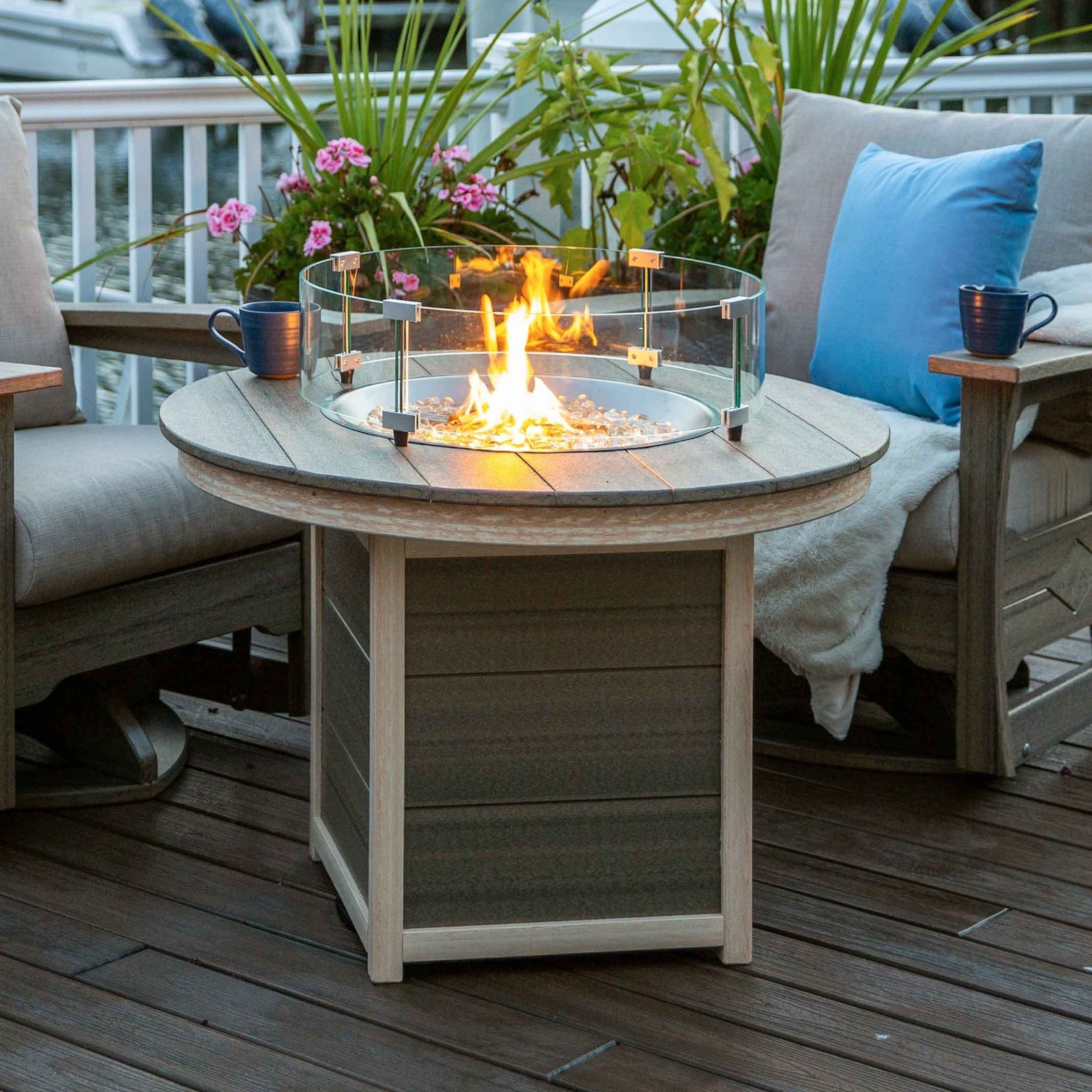 Lit round poly propane fire pit table with glass wind guard and white fire glass, framed by cushioned patio chairs