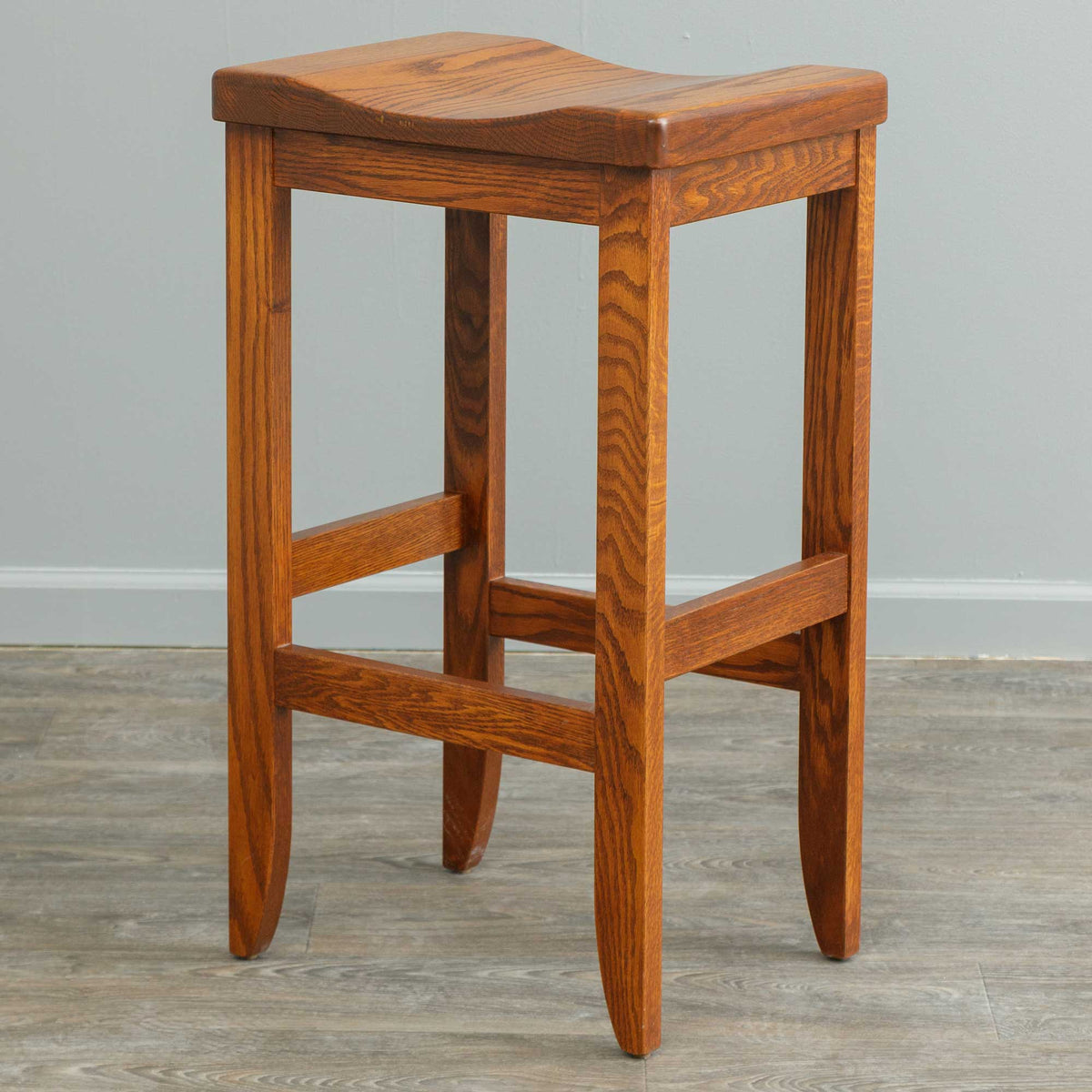 Bar height oak saddle stool with a scooped backless seat, splayed tapered legs and staggered footrest rungs on gray flooring