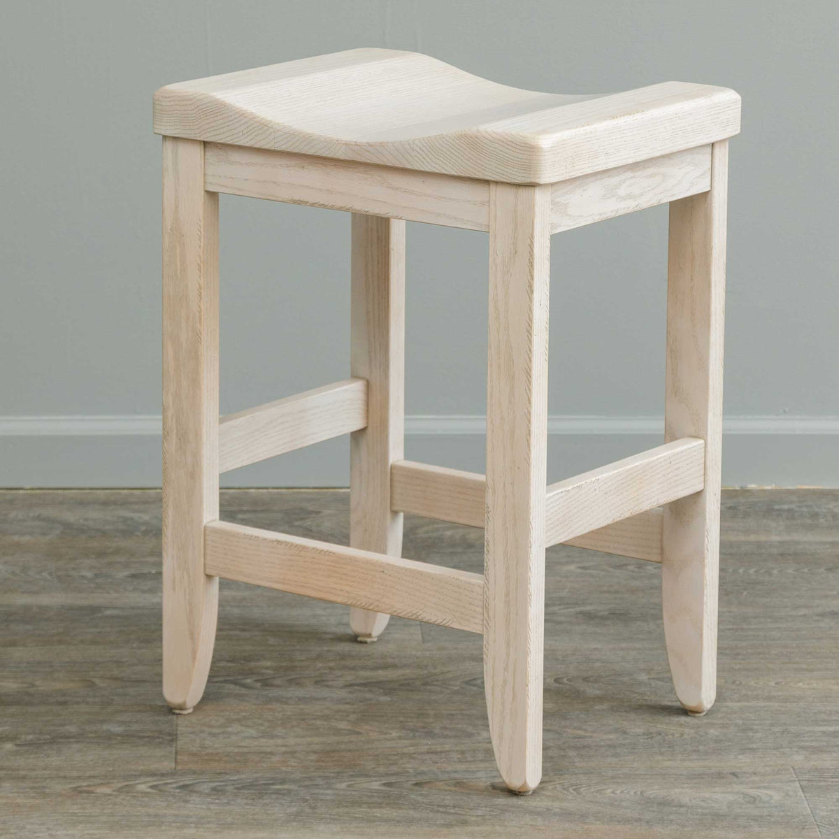 Whitewashed oak saddle bar stool with a contoured backless seat, flared legs and stepped footrest rungs against a gray wall