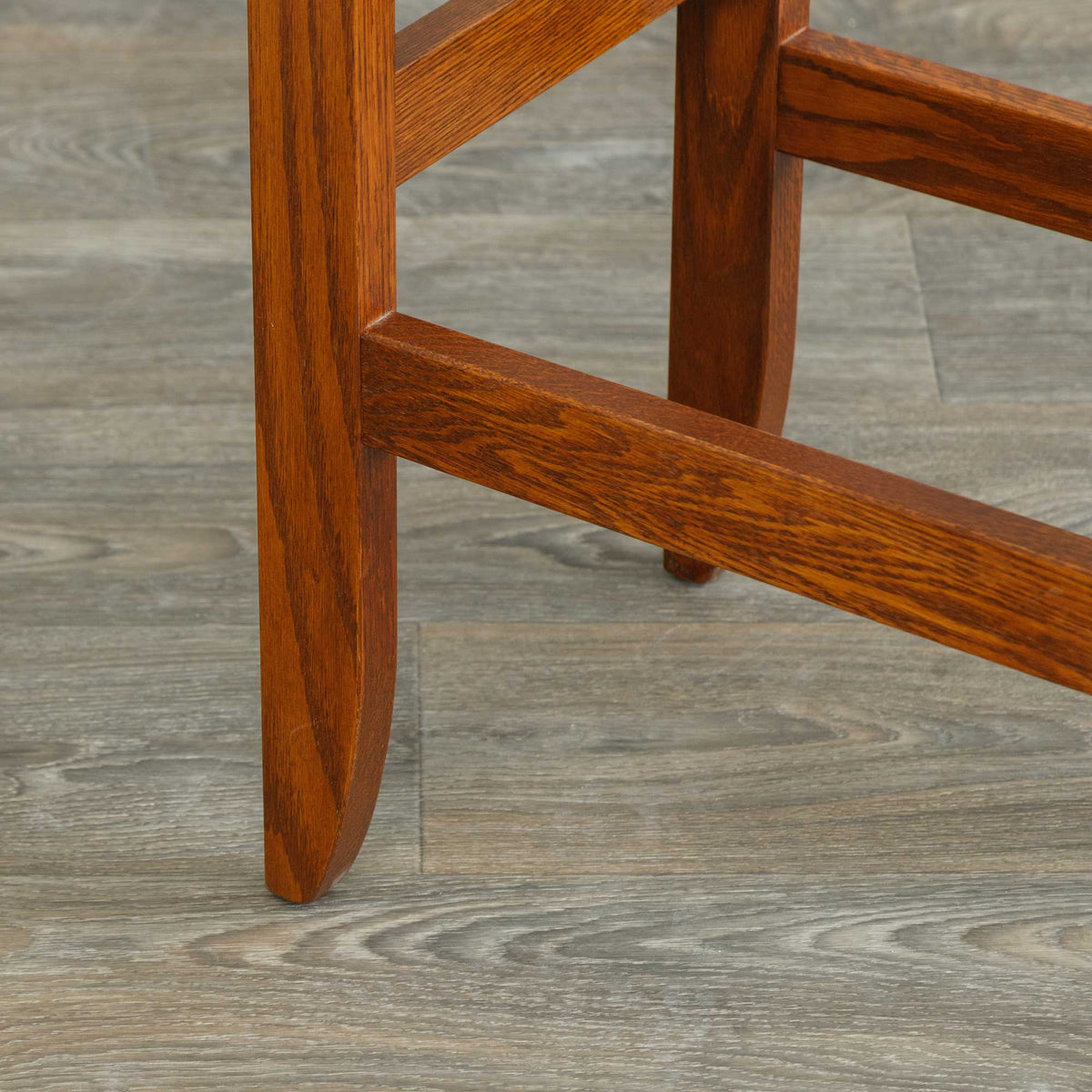 Detail of an oak bar stool leg and footrest rung in a chestnut stain, resting on gray plank flooring