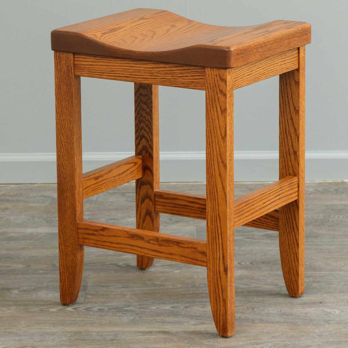 Golden oak backless bar stool with a curved saddle seat, tapered legs and sturdy footrest rungs, on gray wood flooring