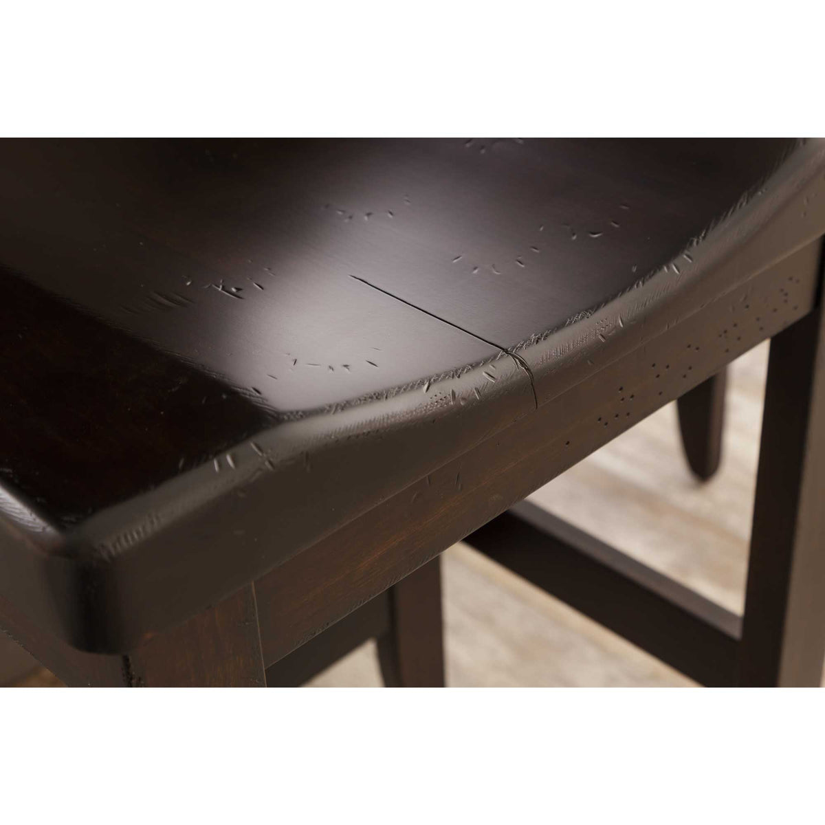 Close-up of a dark espresso saddle stool seat showing distressed marks, curved edge and the apron below