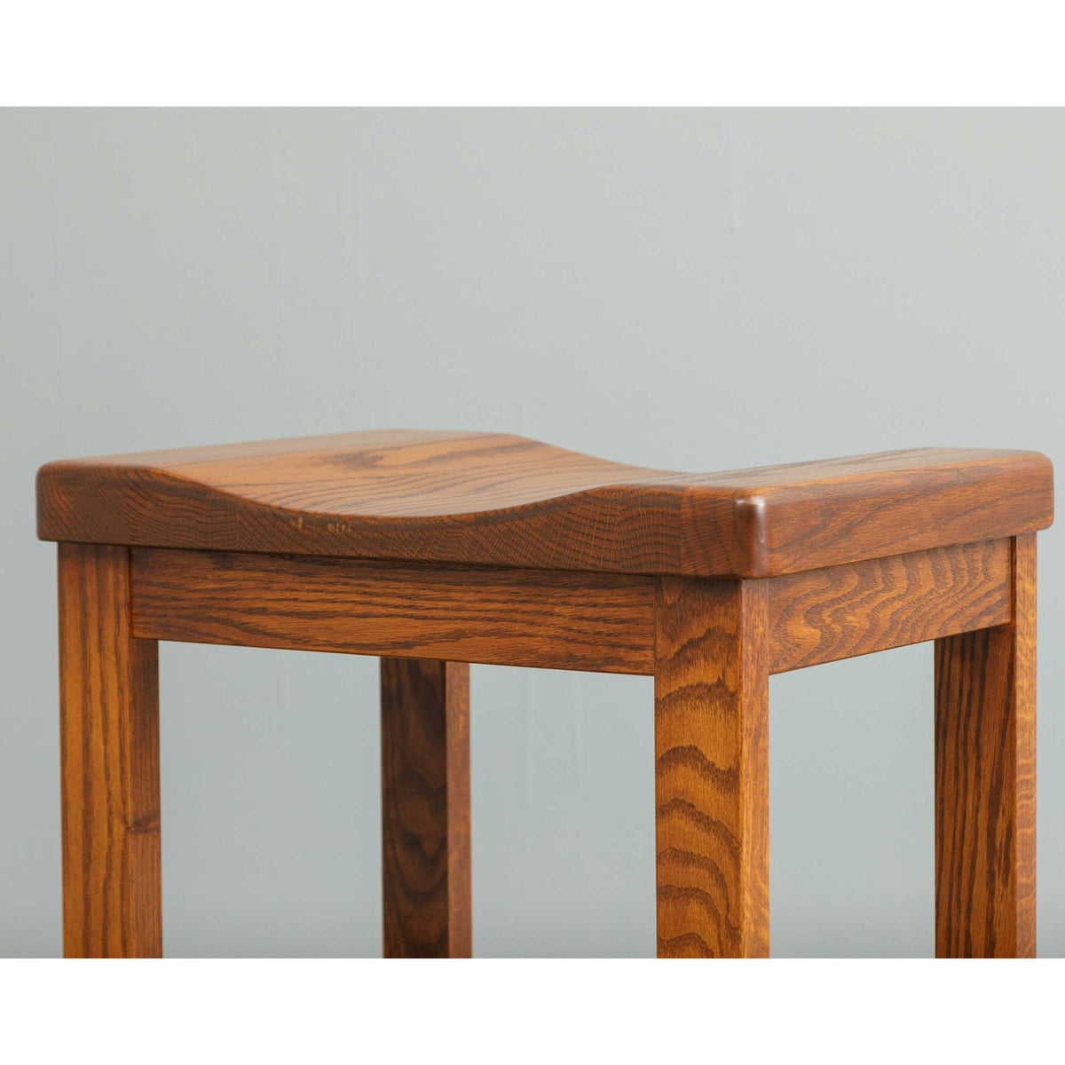 Angled close-up of a quartersawn oak saddle stool seat and leg joint showing the medium brown stain and grain