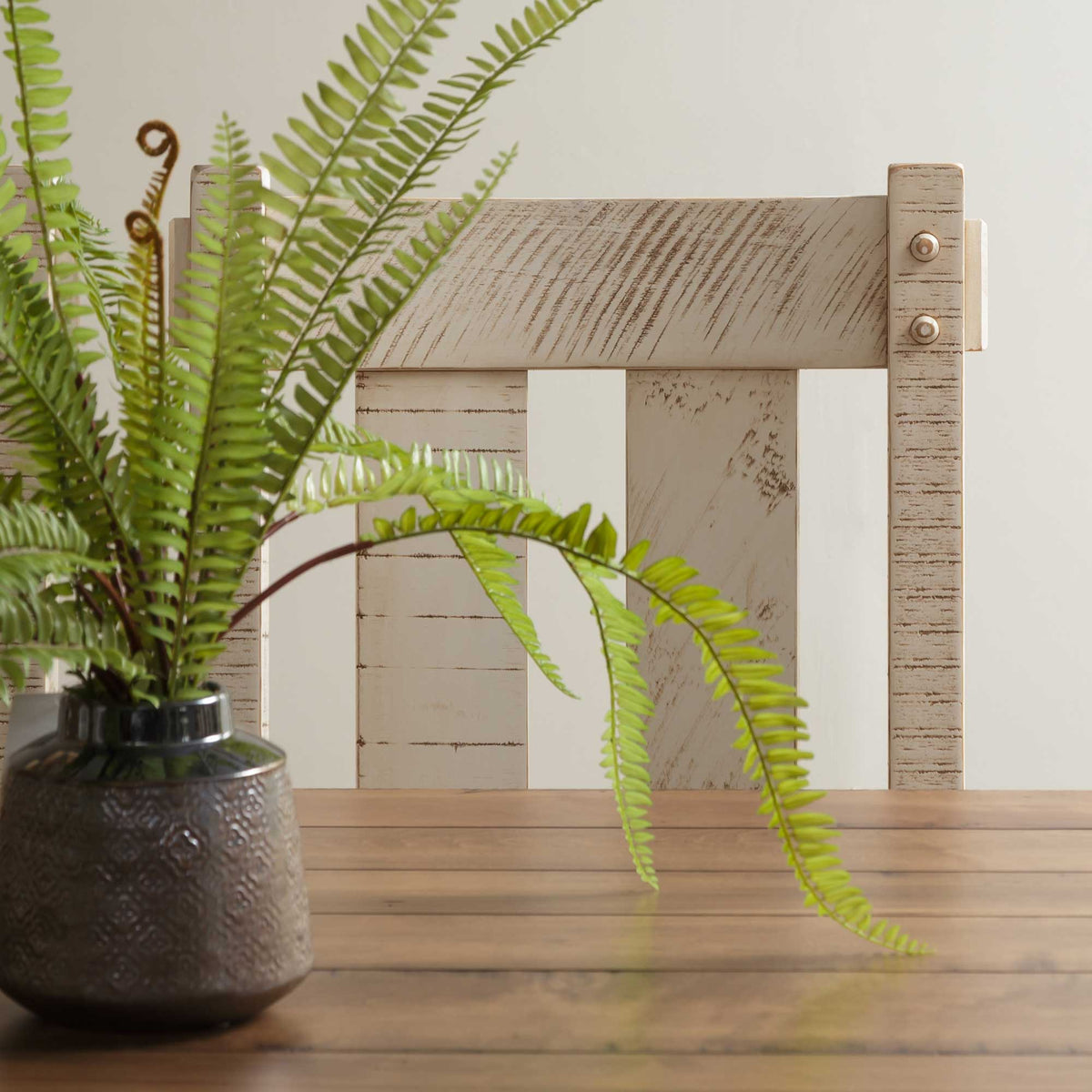 Detail of a distressed white rustic chair back behind a fern in a metal pot on a wood dining table