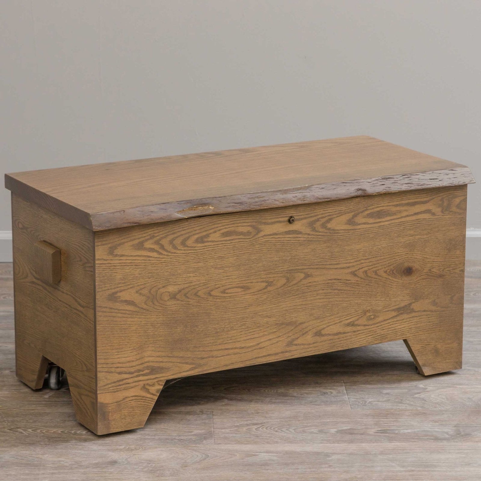 Rustic oak live edge hope chest with a plank lid, side handle and cutout bracket feet on gray flooring