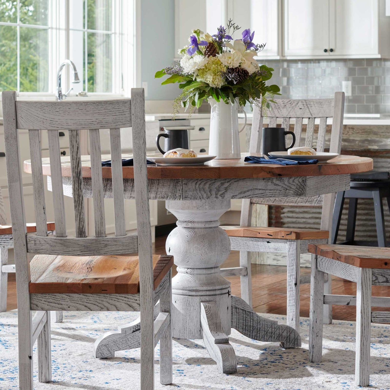 Whitewashed pedestal dining table with warm plank top styled with slat-back chairs in a white farmhouse kitchen