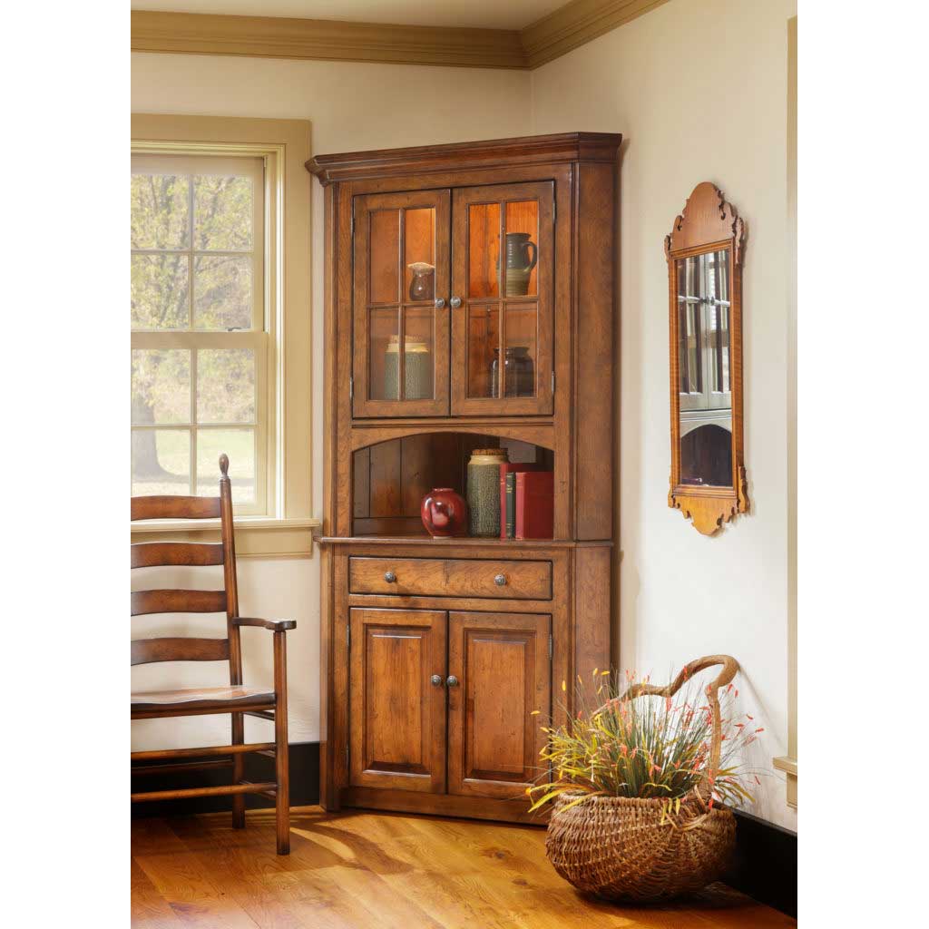 Cherry corner china cabinet with two glass mullion doors, open display shelf, drawer and two panel doors in a sunlit dining room