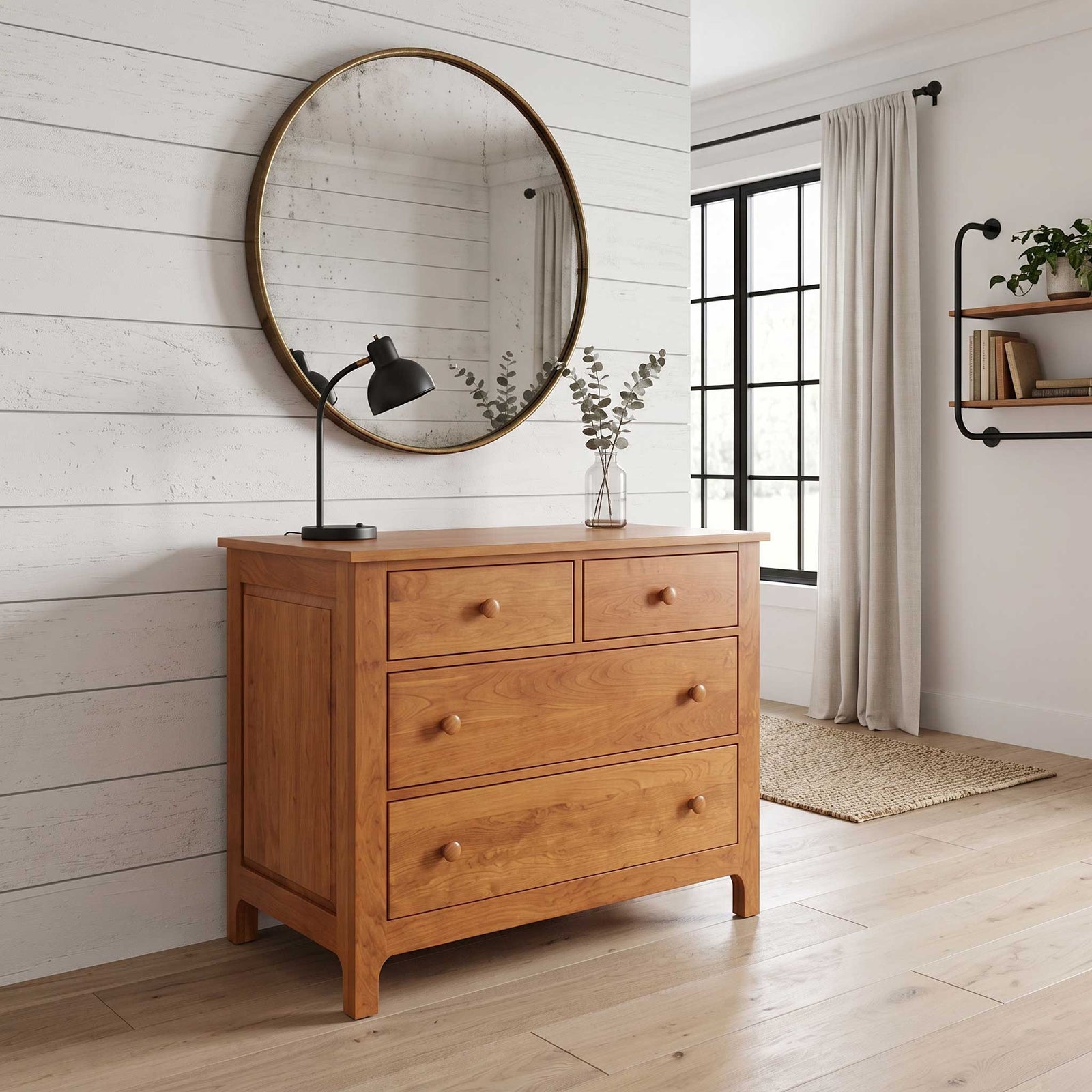A warm medium-brown wooden dresser with three drawers and round knobs, styled in a bright room with a large round mirror, black desk lamp, and a vase on top.