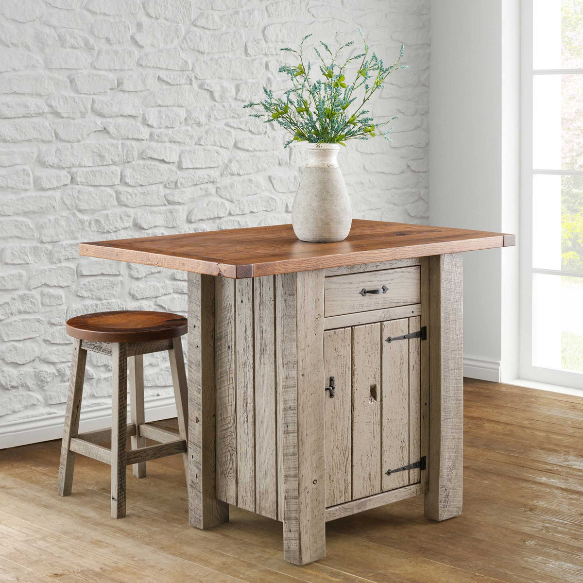 A small rustic kitchen island made of oak wood with a single door, accompanied by two round stools. The island has a light Asbury finish with a van Dyke brown glaze, and it is positioned on a hardwood floor against a white brick wall background.