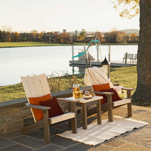 Two-tone taupe modern Adirondack chairs with orange pillows and a square side table on a flagstone patio by a pond