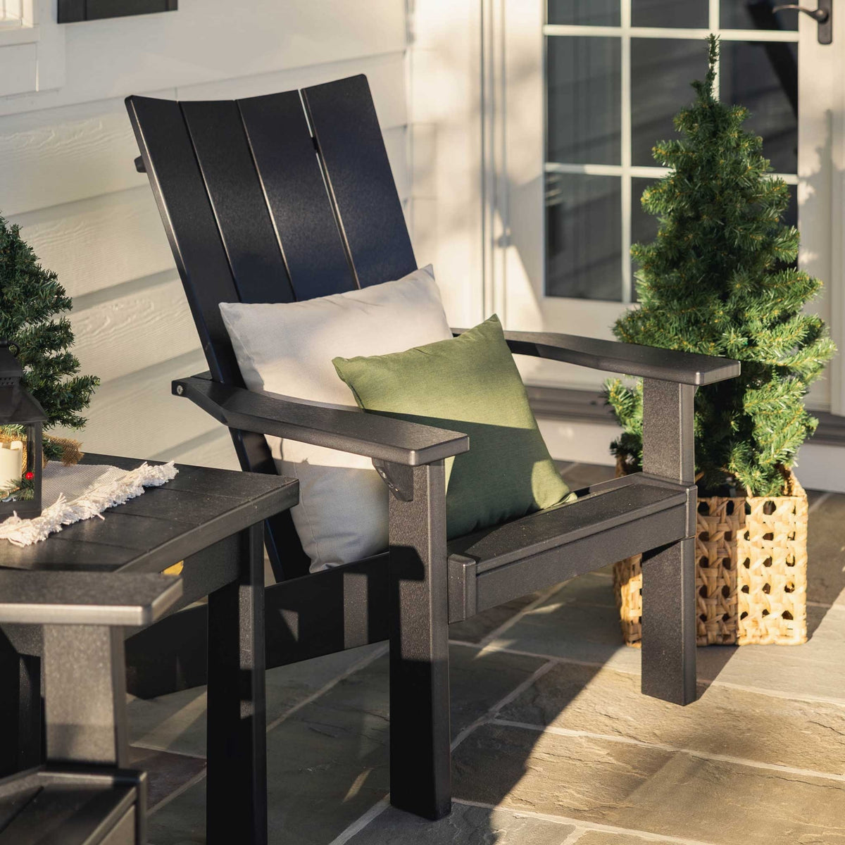 Black contemporary Adirondack chair with cream and green pillows beside a square side table and potted evergreens