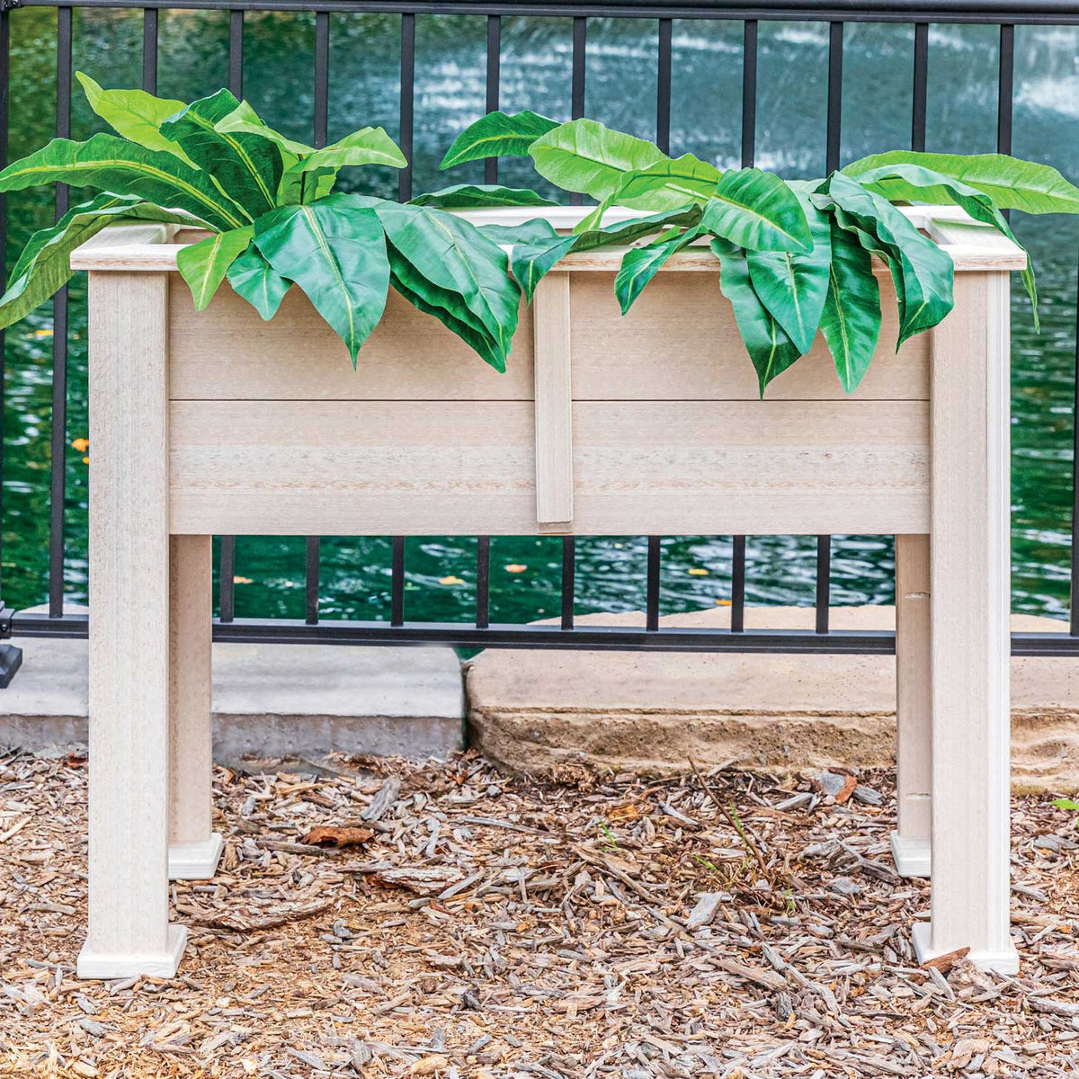 Raised birch poly planter on legs filled with large green leafy plants beside a pond railing