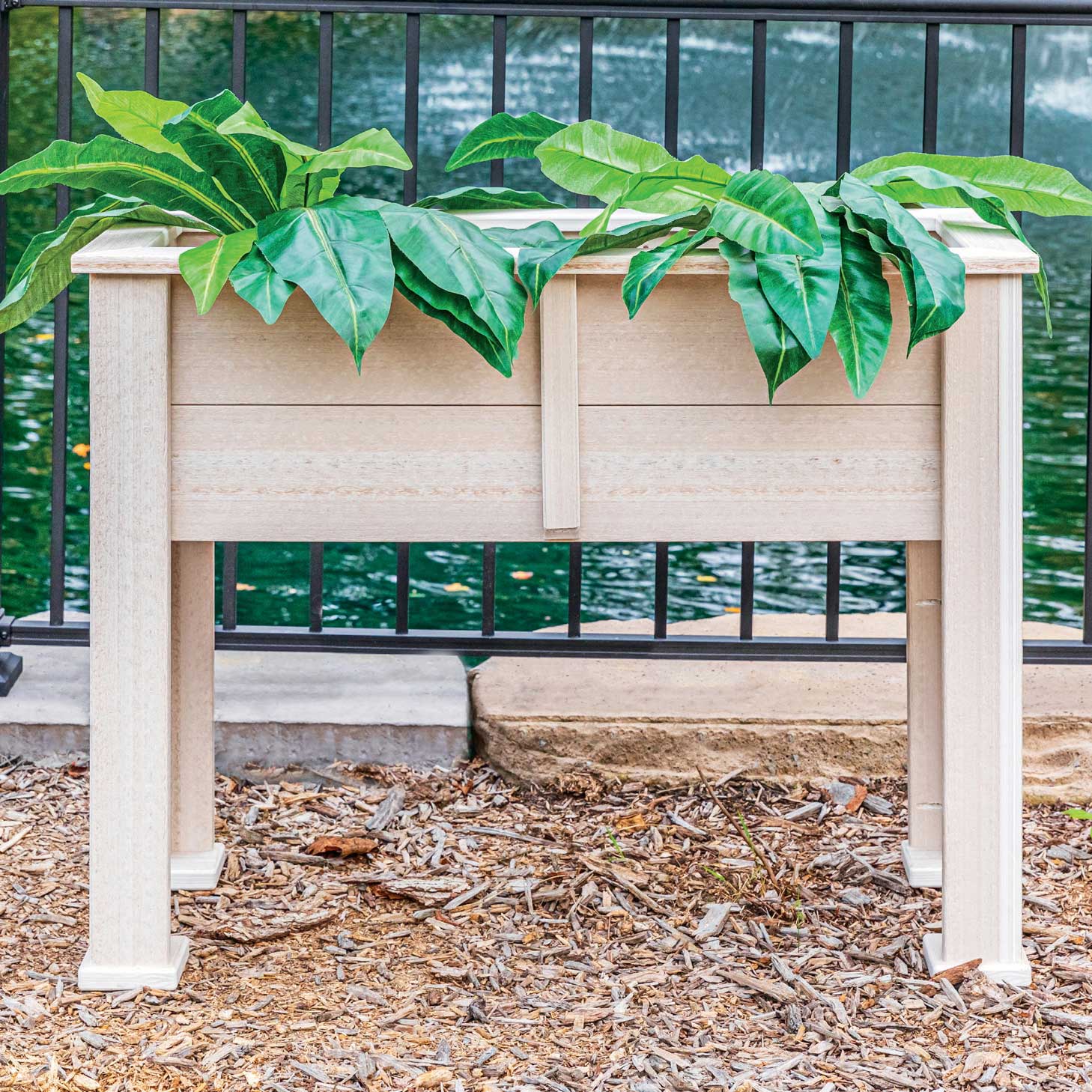 Raised birch poly planter on legs filled with large green leafy plants beside a pond railing