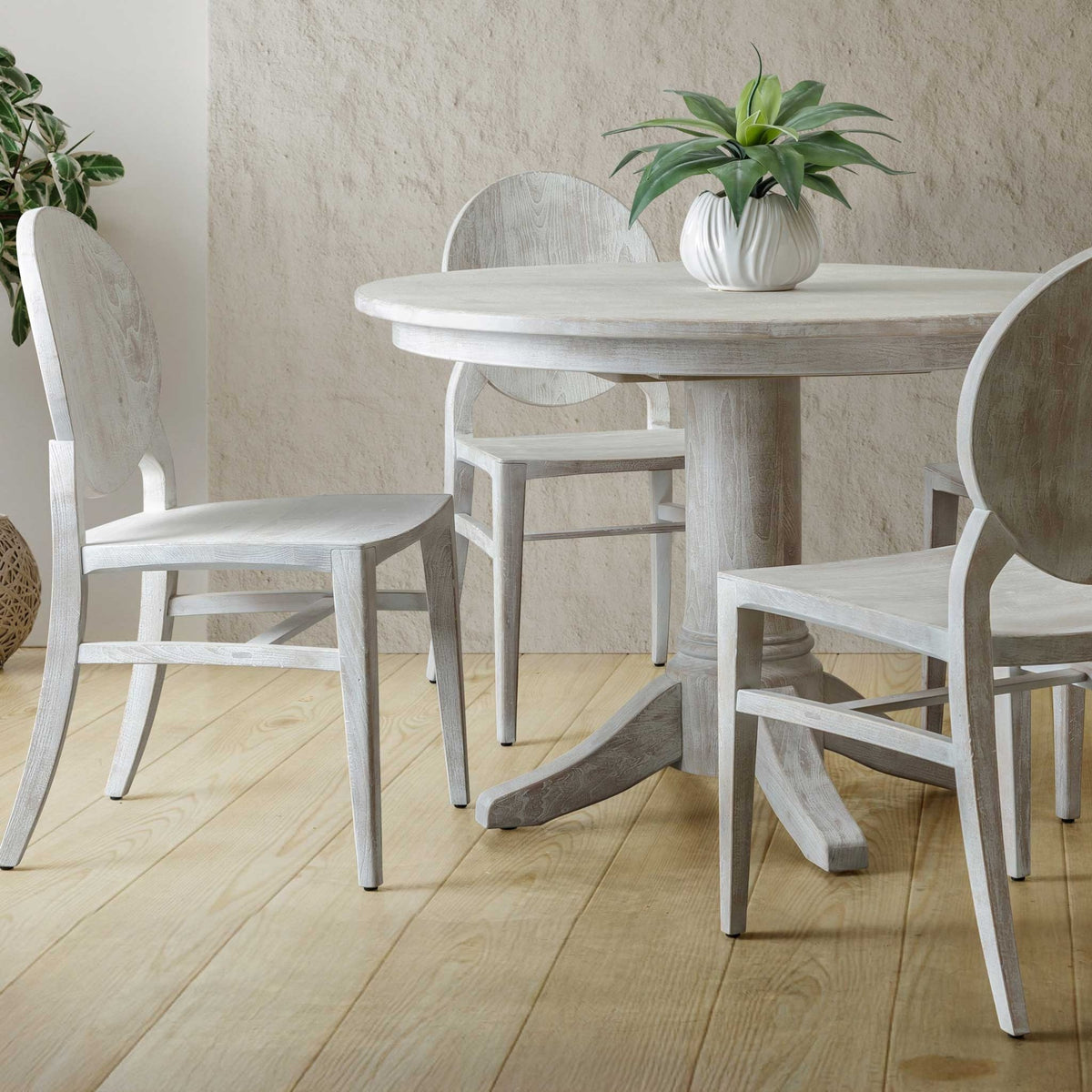 Whitewashed oval back dining chairs with solid wood seats around a round white pedestal table in a bright minimal dining space